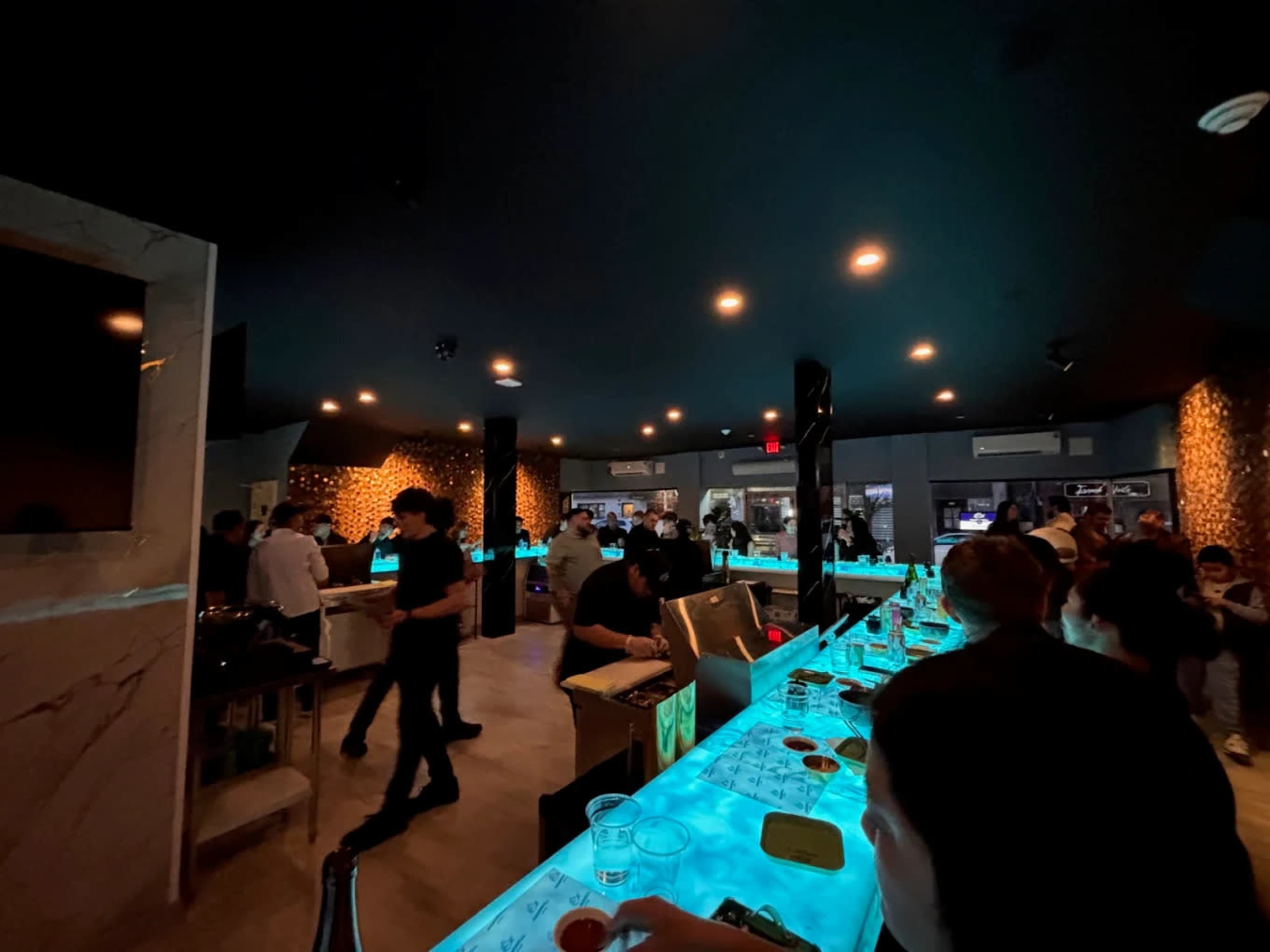 The image shows a dimly lit bar with a blue illuminated countertop, where patrons are gathered, and staff are serving drinks.