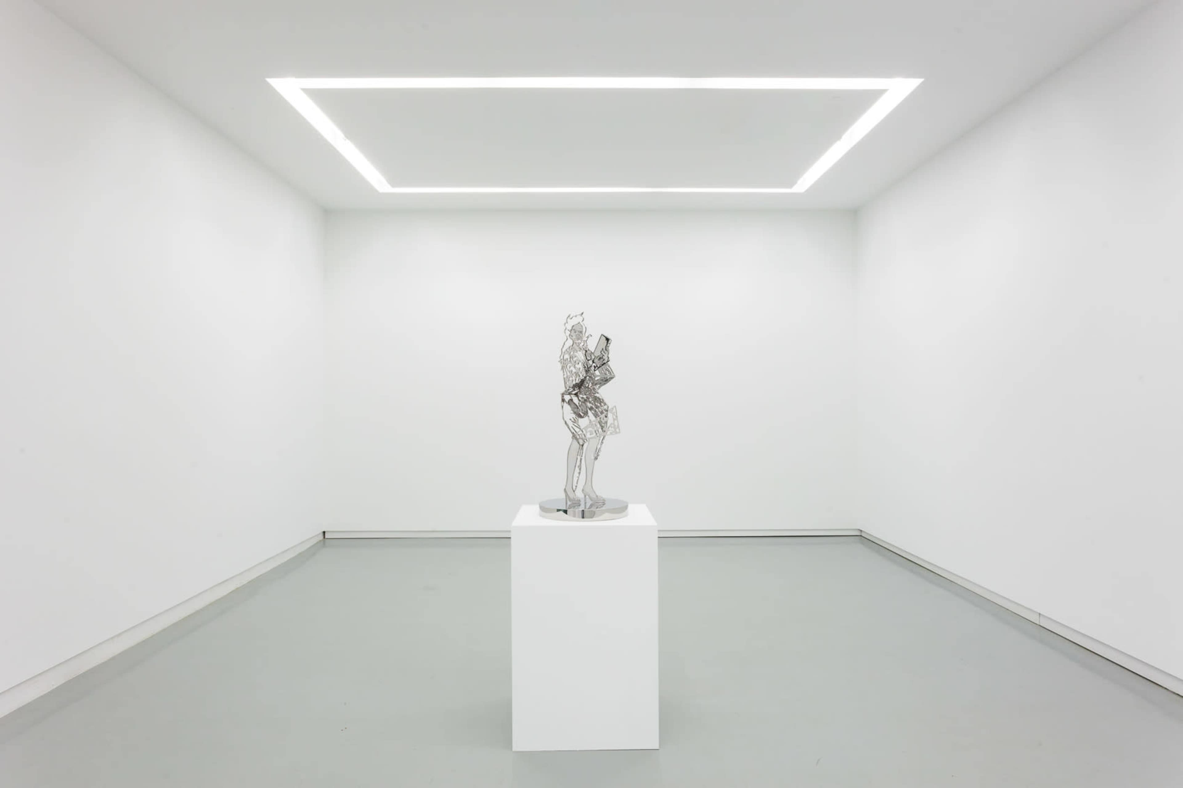 A metallic sculpture stands on a white pedestal in a minimally designed gallery with white walls and a rectangular light fixture.