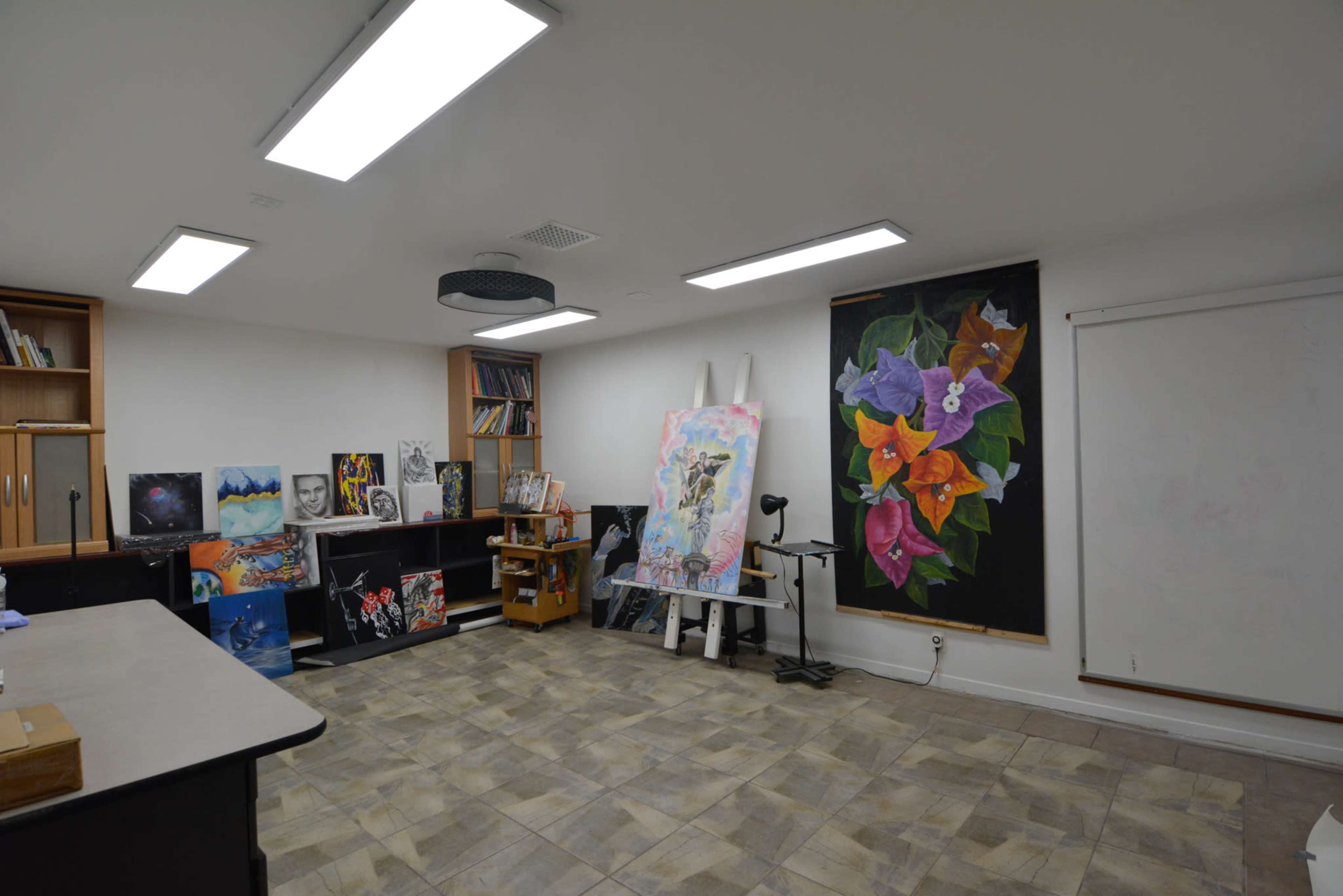 An art studio with several canvases displaying various artworks, organized shelves, and bright overhead lighting.