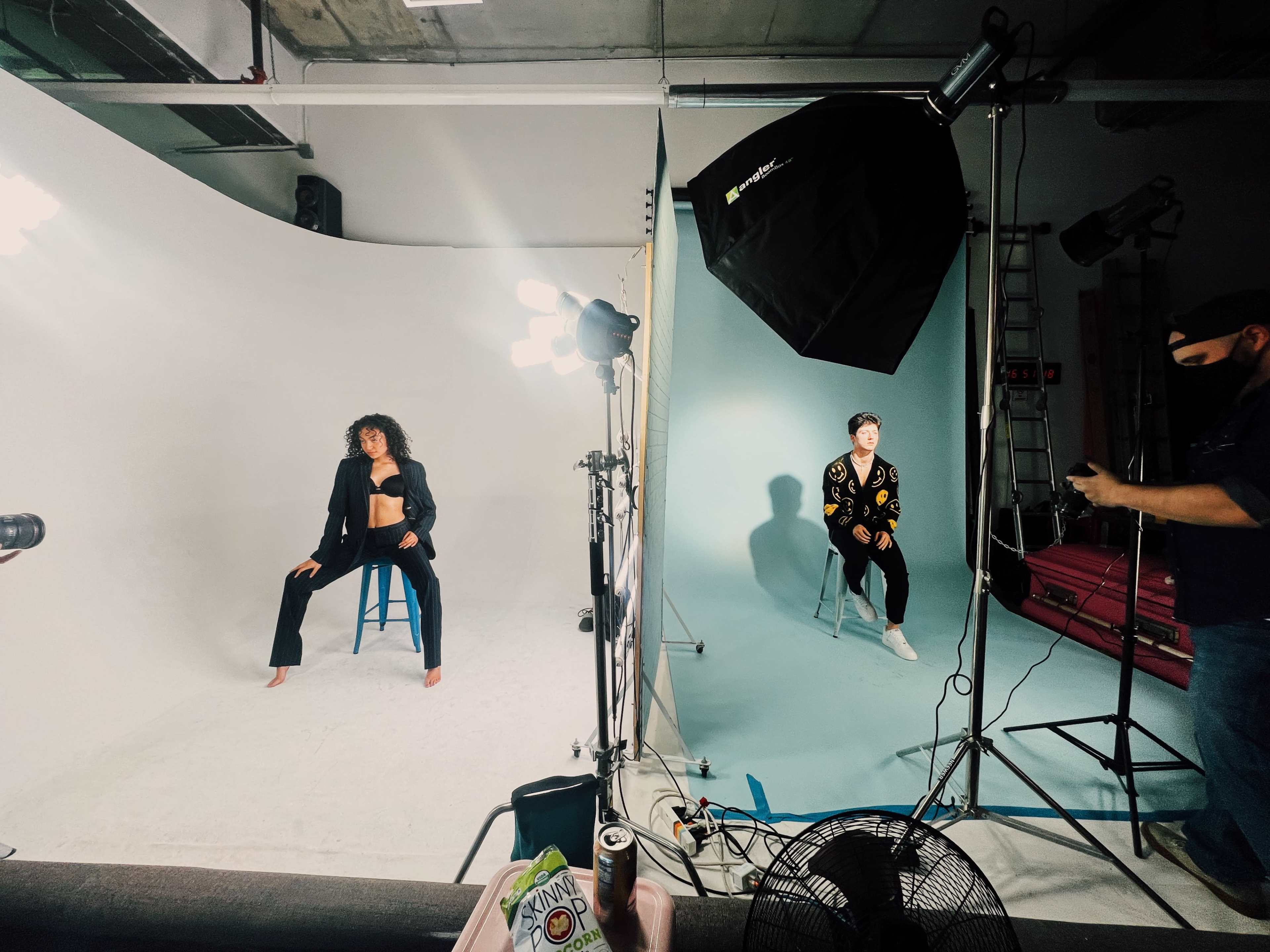 The image shows a photography studio with two models on stools, one dressed in a black outfit and the other in a colorful sweater, while a photographer adjusts lighting equipment.