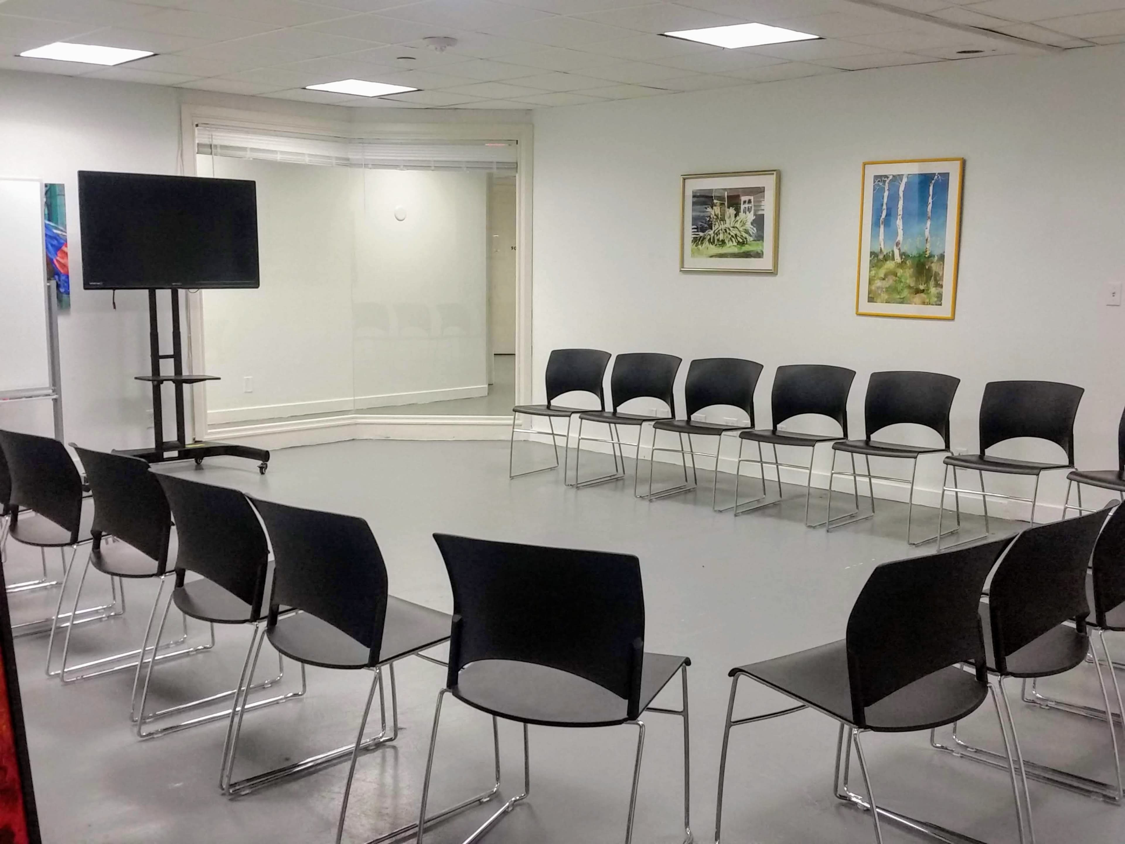 A room with a circular arrangement of black chairs and a television screen on a stand.