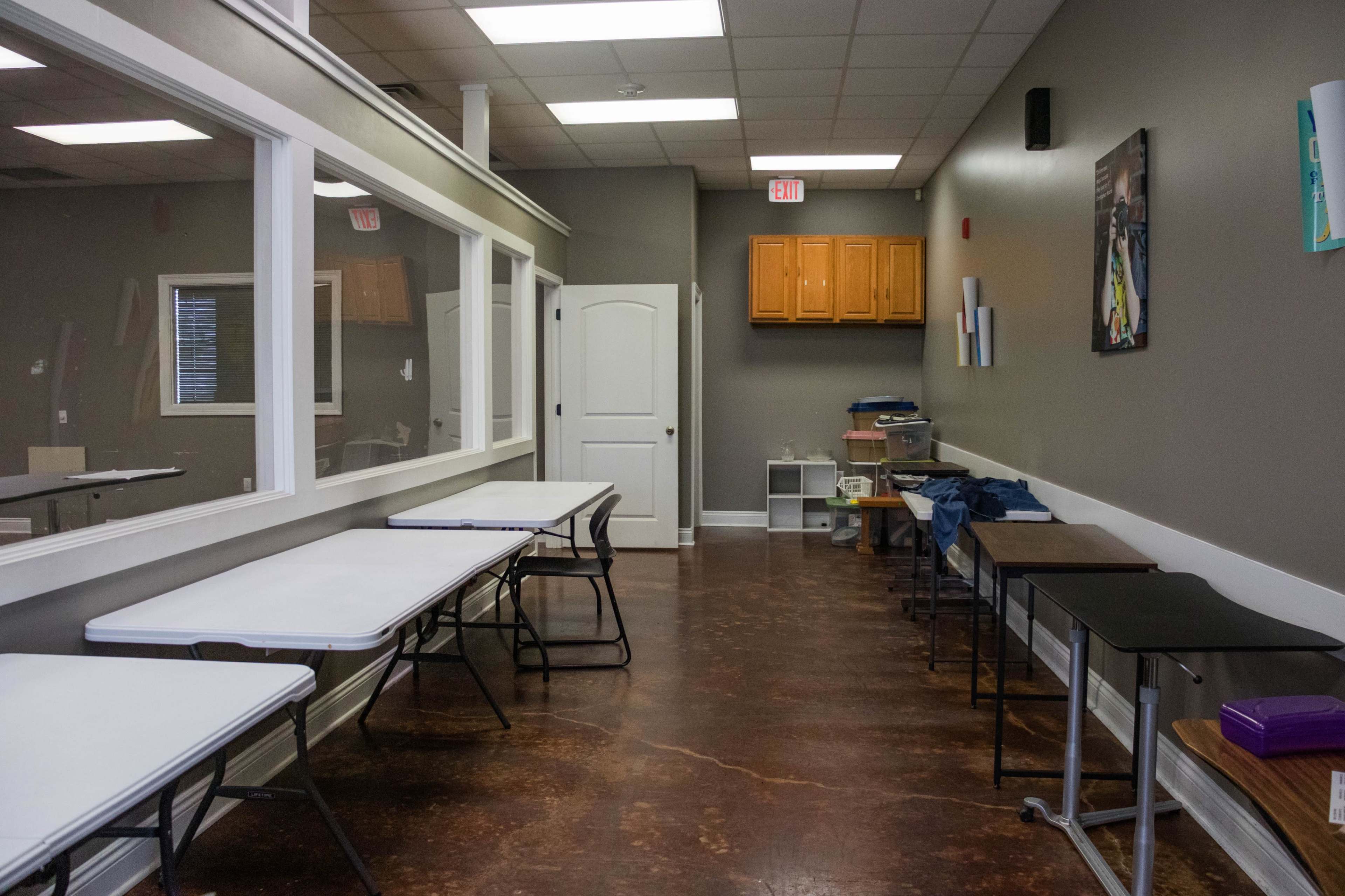 The image shows a spacious room with multiple folding tables arranged on either side and a set of wooden cabinets mounted on the wall.