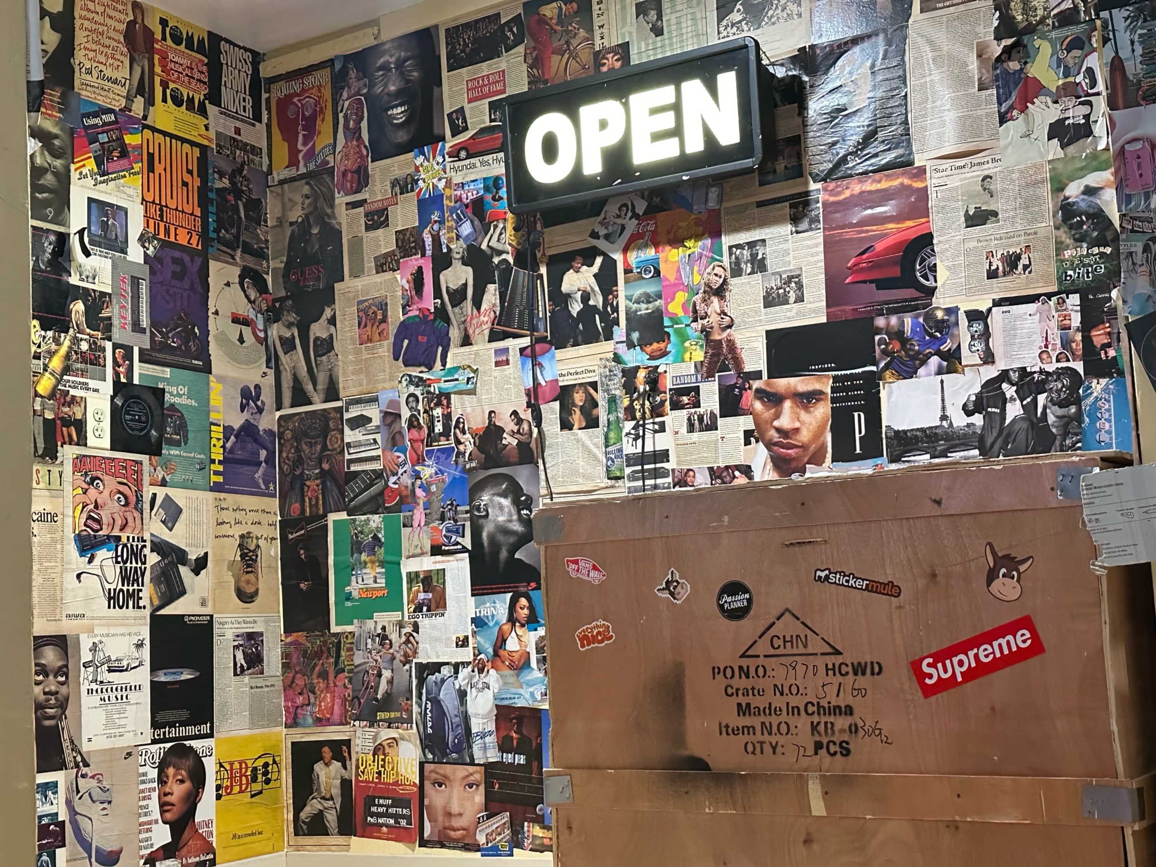 A wall adorned with various posters and magazine clippings, featuring an illuminated "OPEN" sign above a wooden crate covered in stickers.