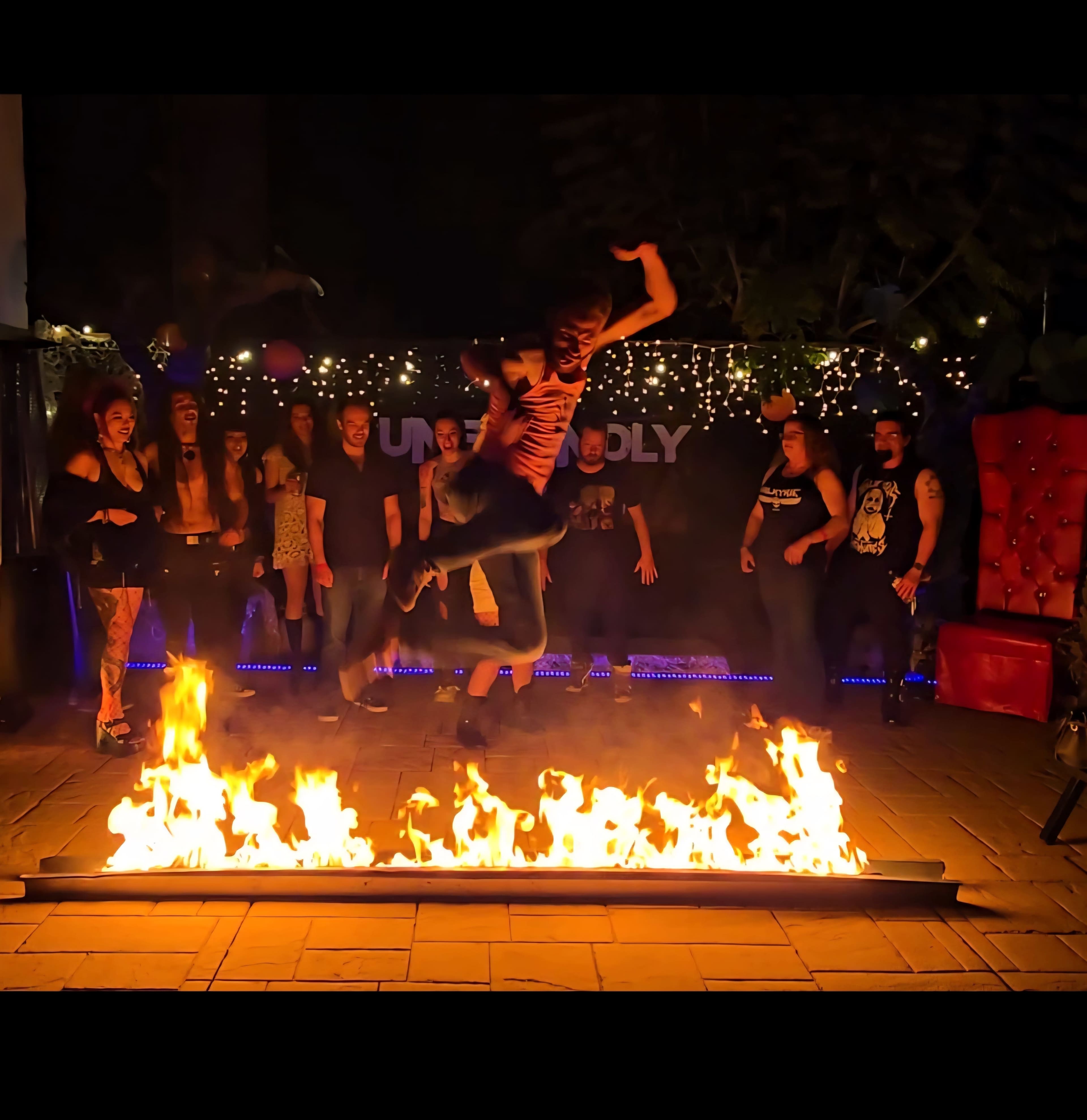 A person leaps over a flaming barrier while a crowd watches in the background at a nighttime event.