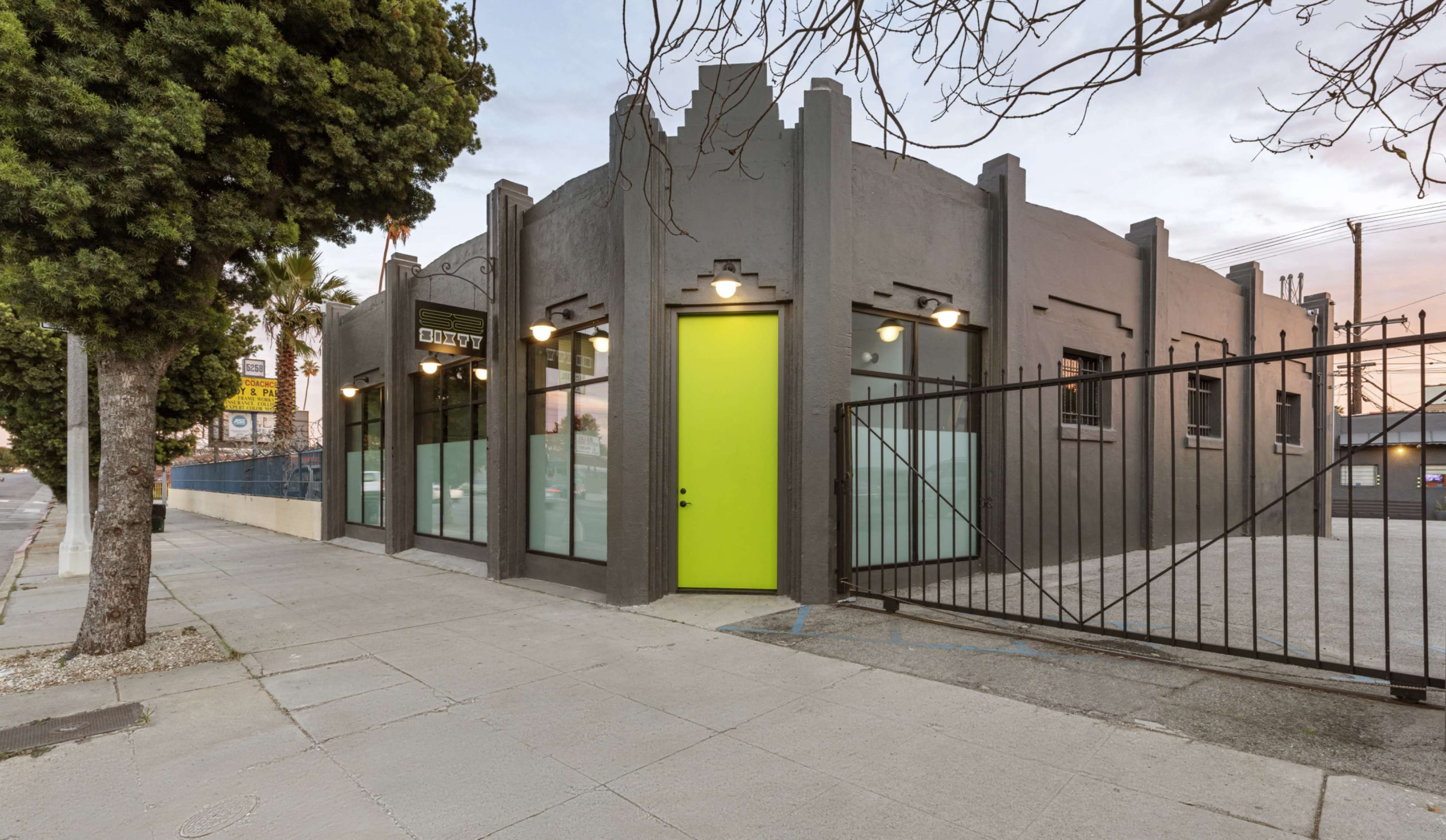 A modern building with a striking green door is situated on a street corner, featuring a gray exterior and large windows, surrounded by a fenced parking area.