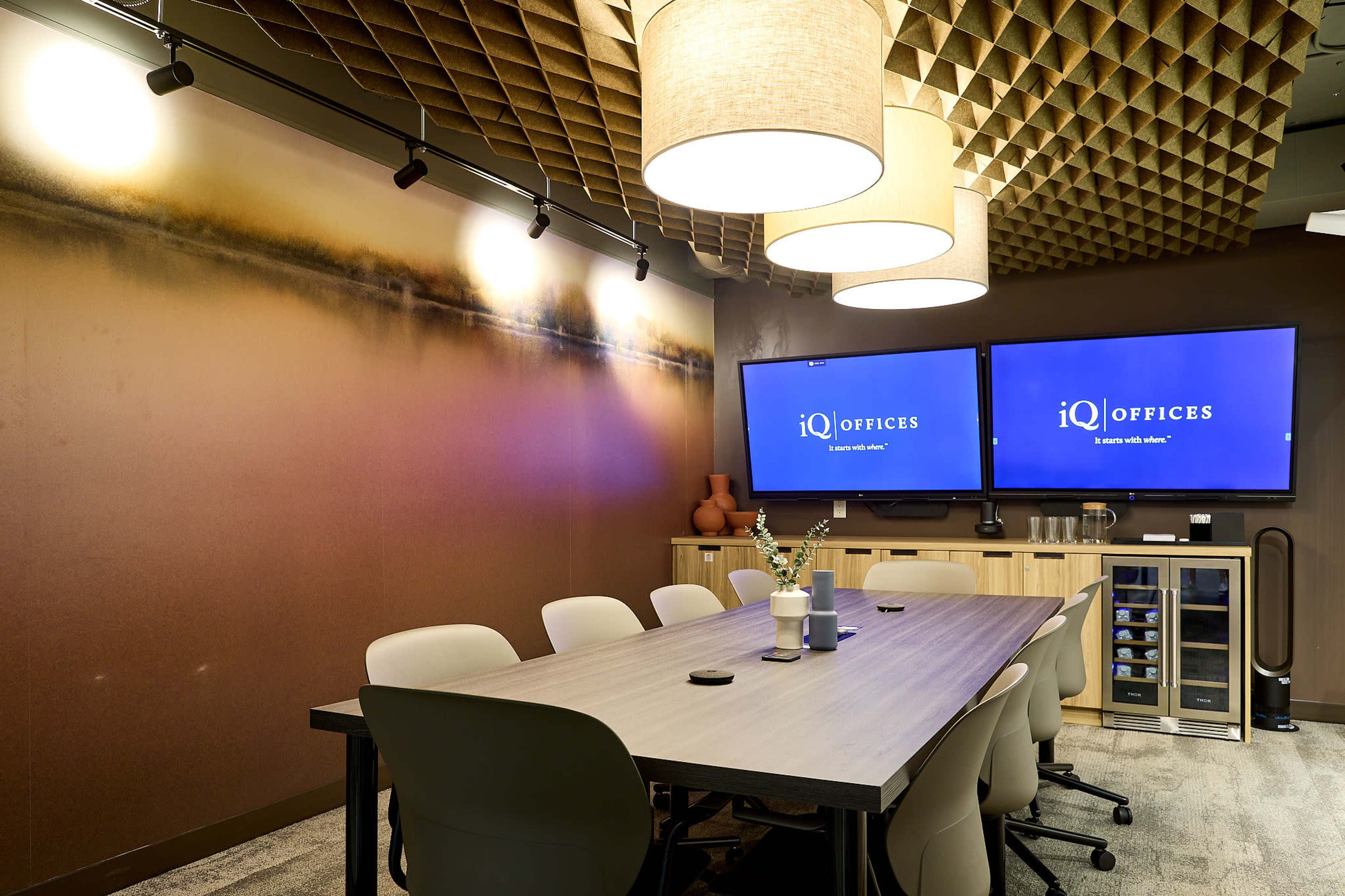 The image shows a modern conference room with a long table surrounded by ergonomic chairs, two large screens mounted on the wall, and stylish overhead lighting.
