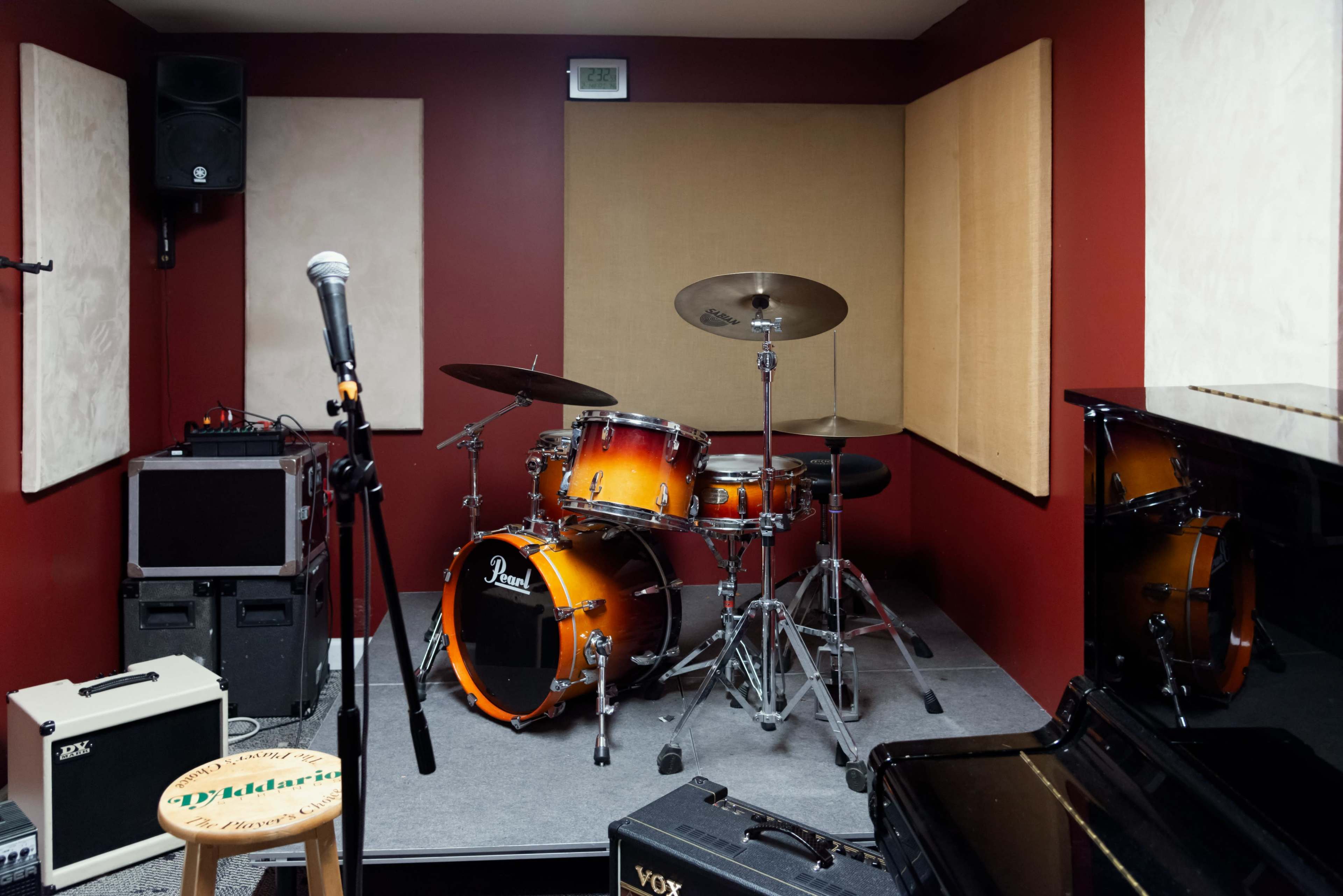 The image shows a small music rehearsal space featuring a drum set, microphone stand, and various audio equipment against red walls.