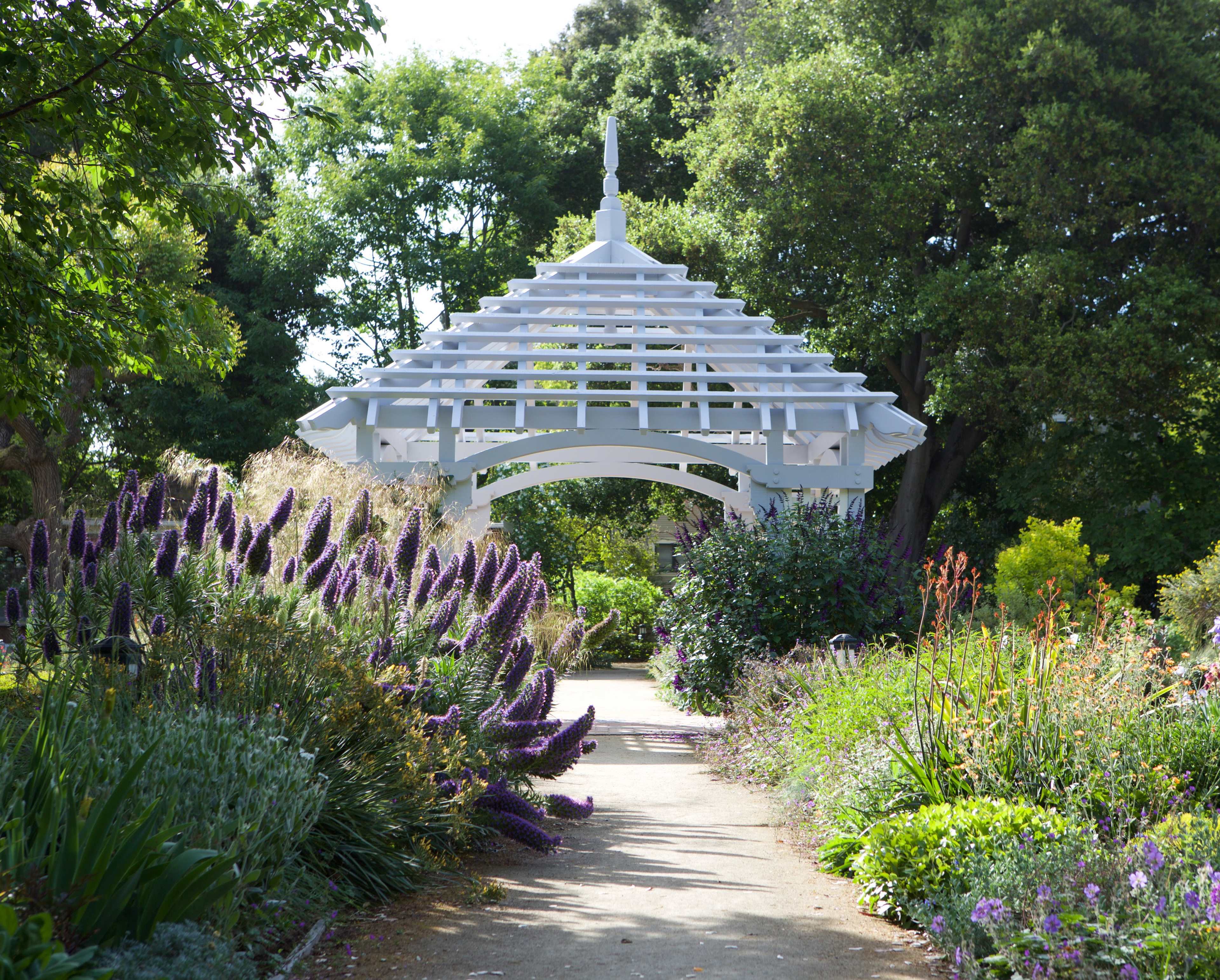 A white wooden gazebo is situated at the end of a pathway lined with colorful flowering plants and lush greenery.