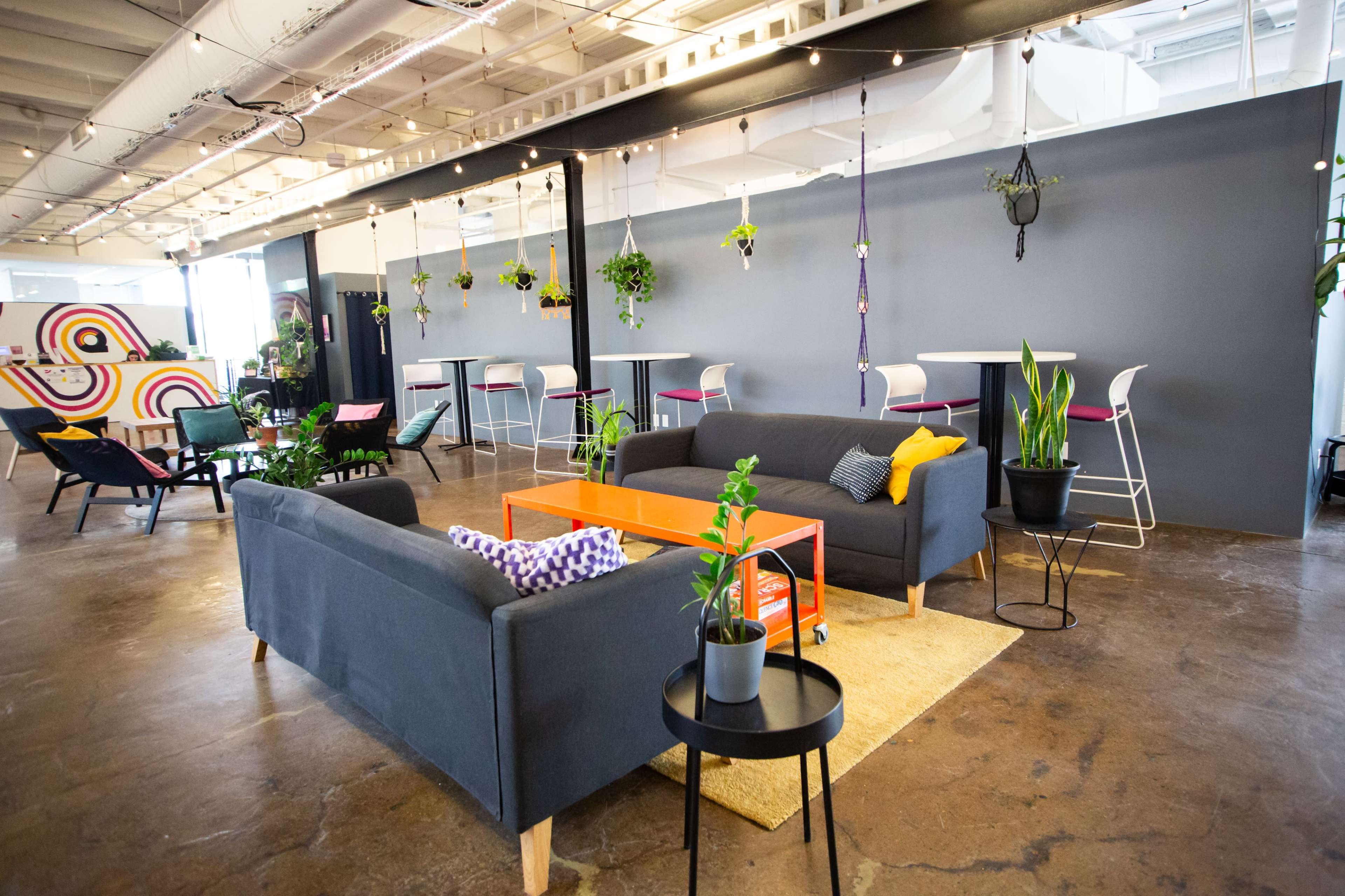 The image shows a modern office space with lounge seating, a communal area featuring plants, and a mix of colorful furniture.