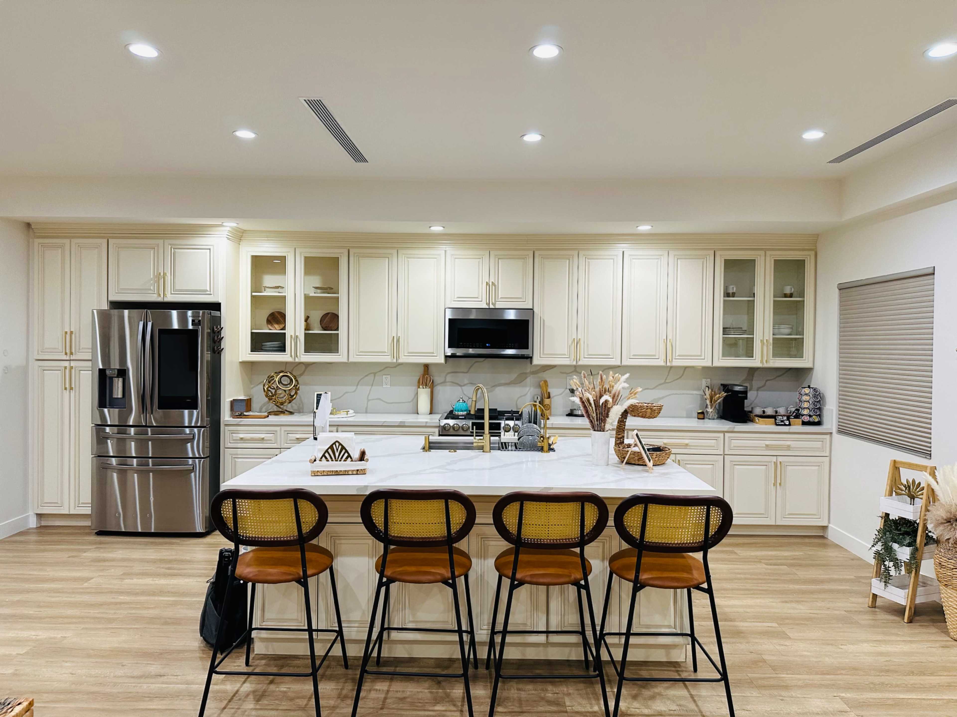 A modern kitchen features white cabinetry, a large island with bar stools, stainless steel appliances, and a marble countertop.