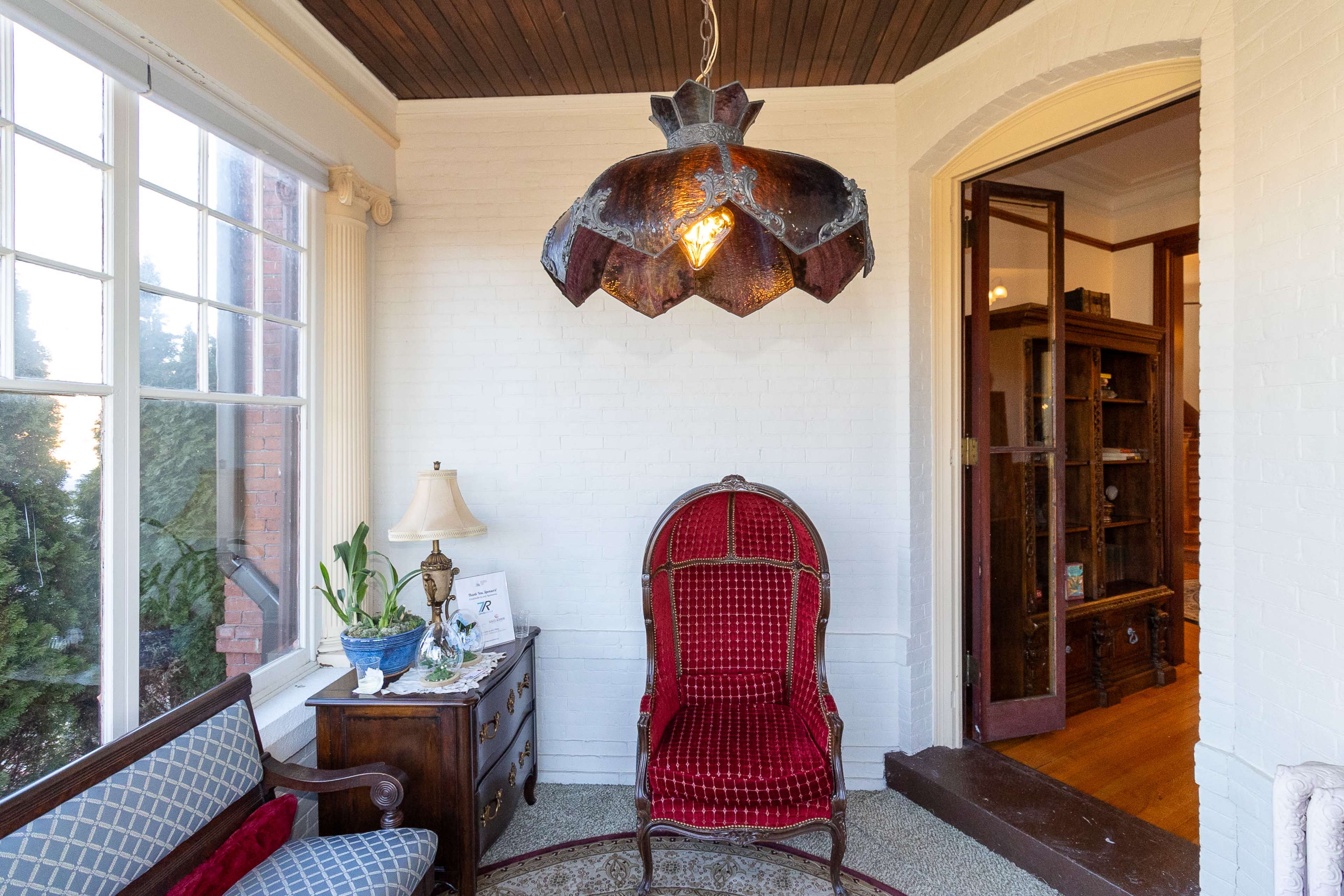A corner of a room featuring a red upholstered armchair beneath a vintage light fixture, a side table with a lamp, and a large window letting in natural light.