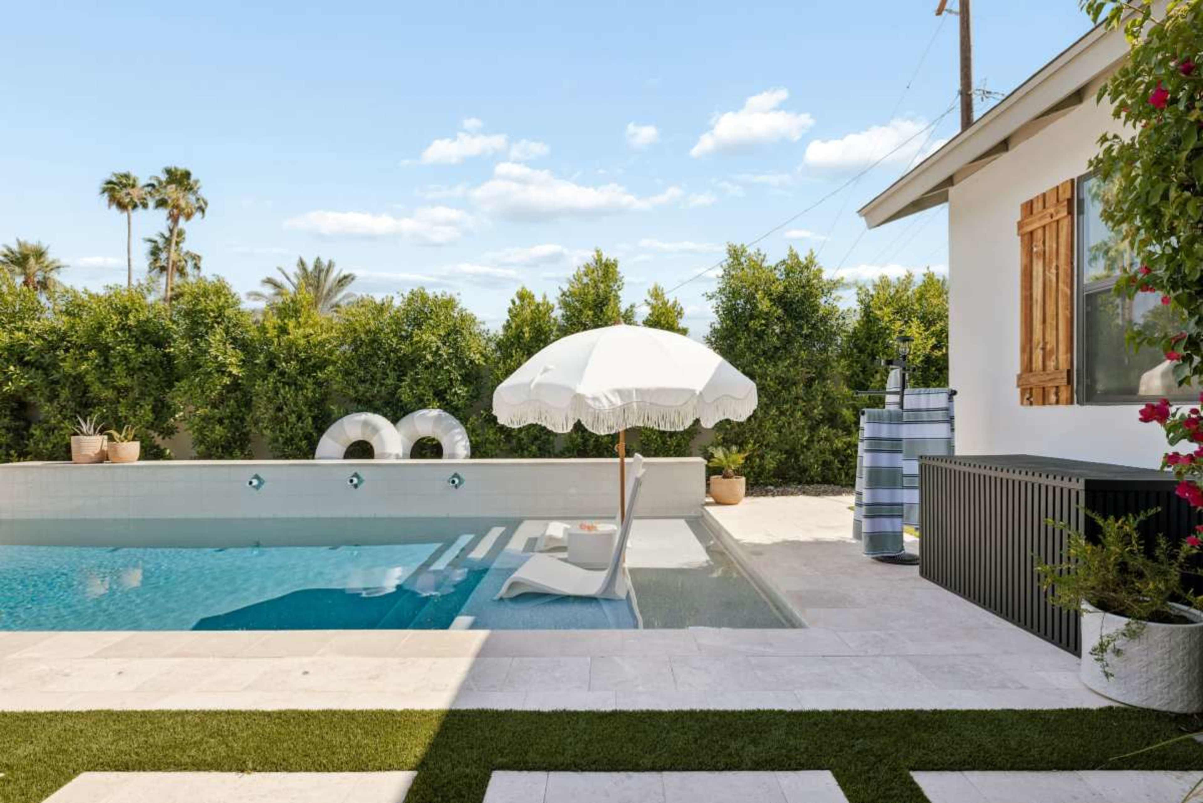 A swimming pool with an umbrella and lounge chair is surrounded by lush greenery and a modern house.