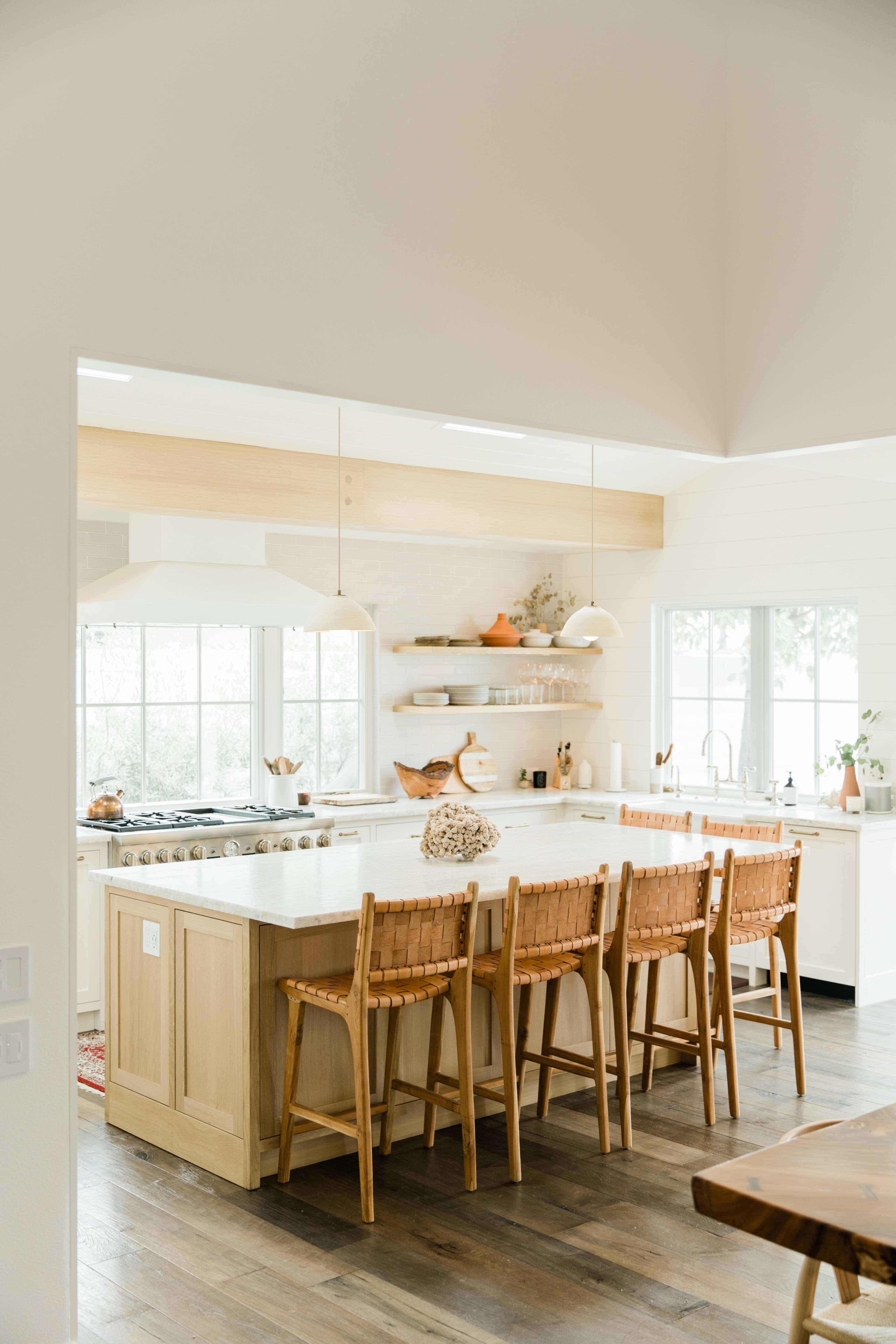 A modern kitchen with a large island, wooden bar stools, and a white countertop under natural light from windows.