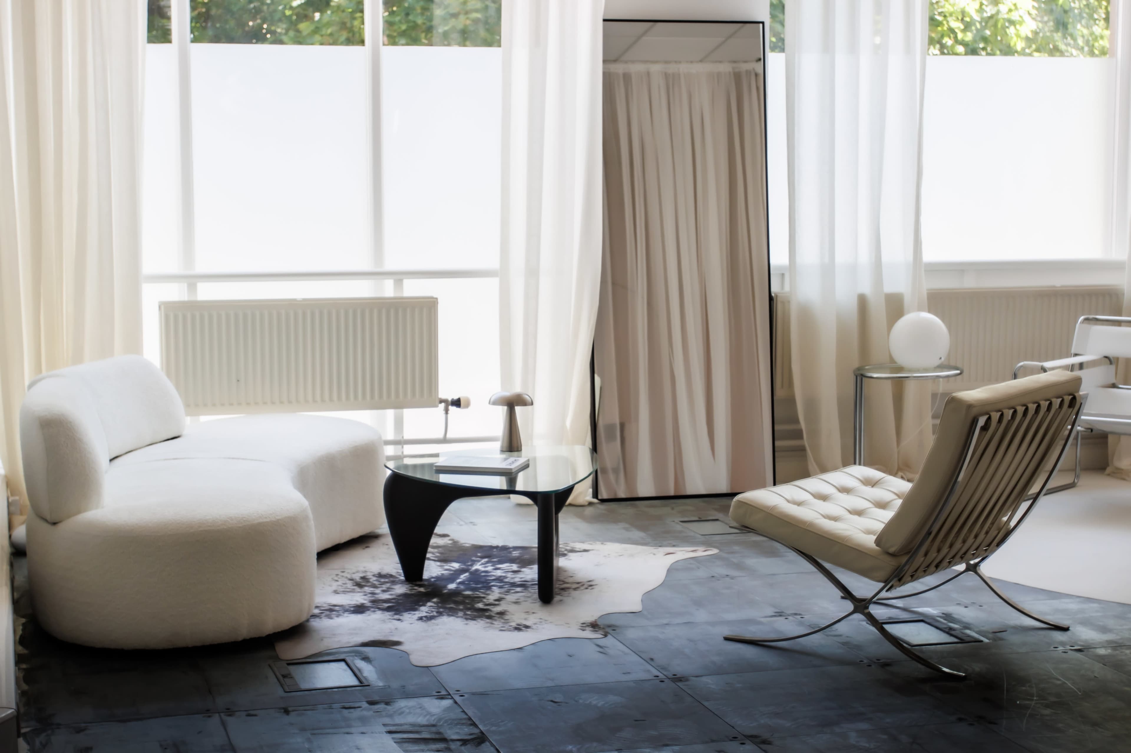 A bright, modern living room featuring a white curved sofa, a glass coffee table, and a minimalist chair on a dark floor with sheer curtains allowing natural light.