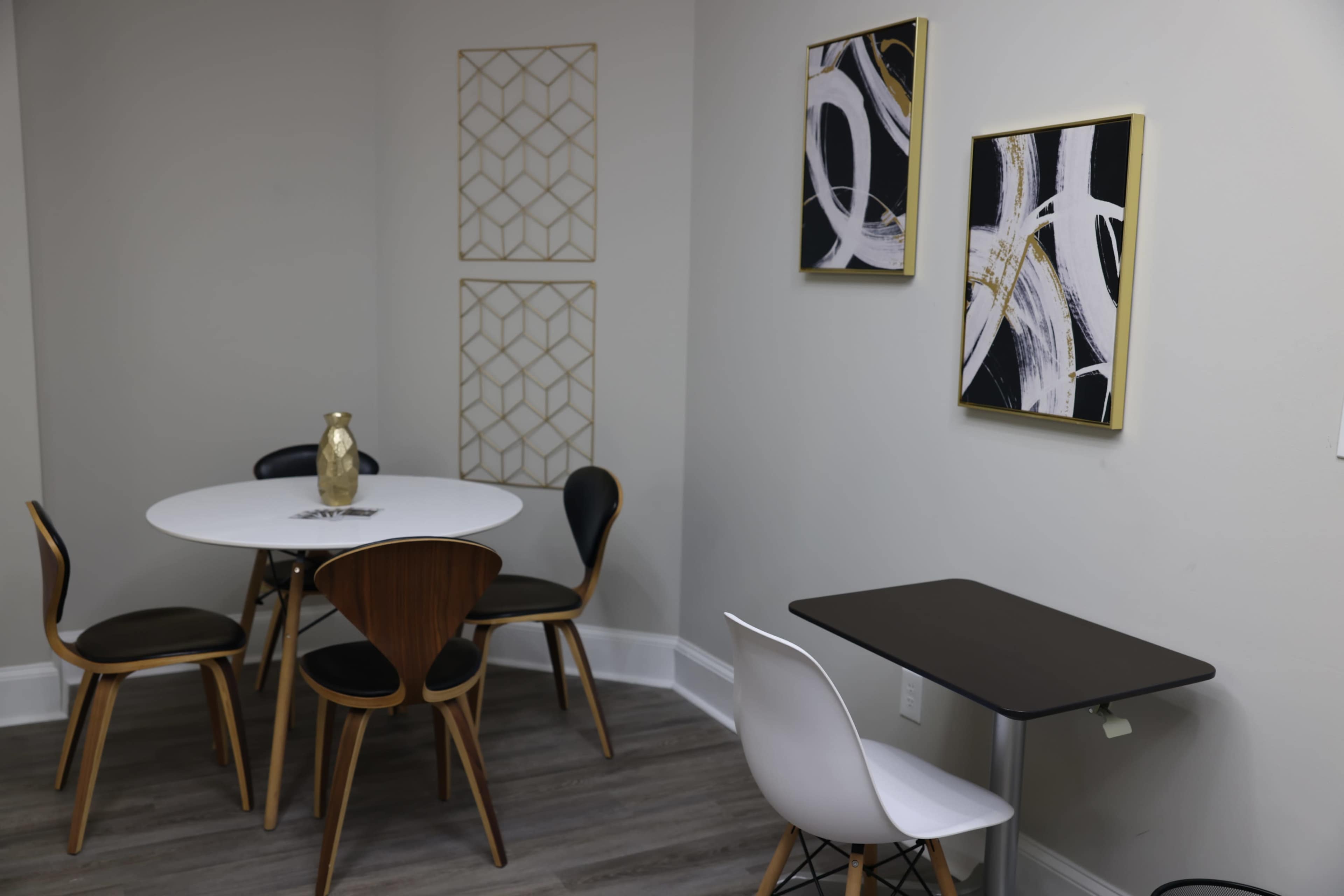 A minimalist dining area features a round white table surrounded by black and wooden chairs, with two abstract wall art pieces and geometric wall decor.