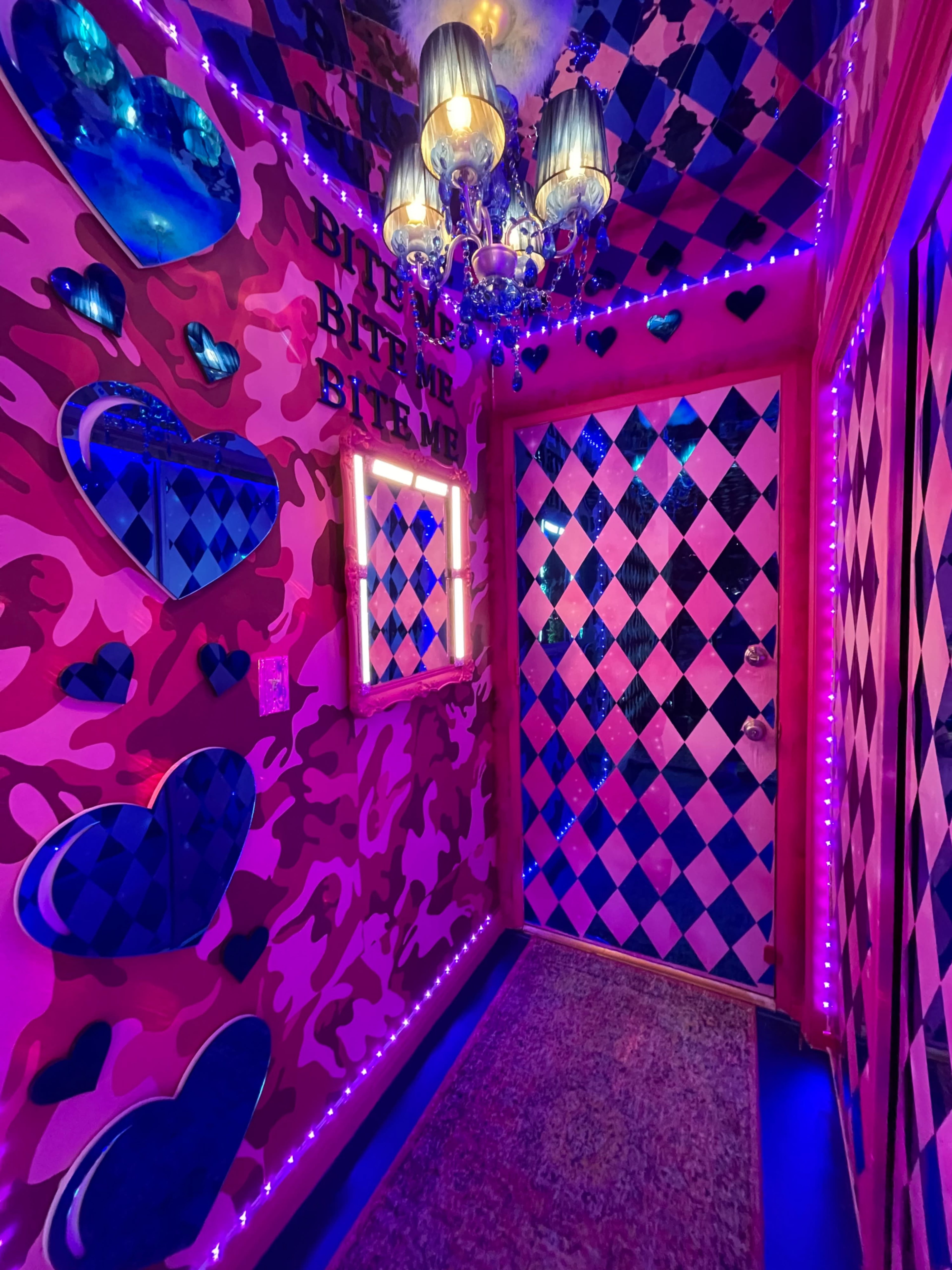 A brightly decorated hallway with pink camouflage walls, heart-shaped mirrors, a checkered door, and purple LED lights.
