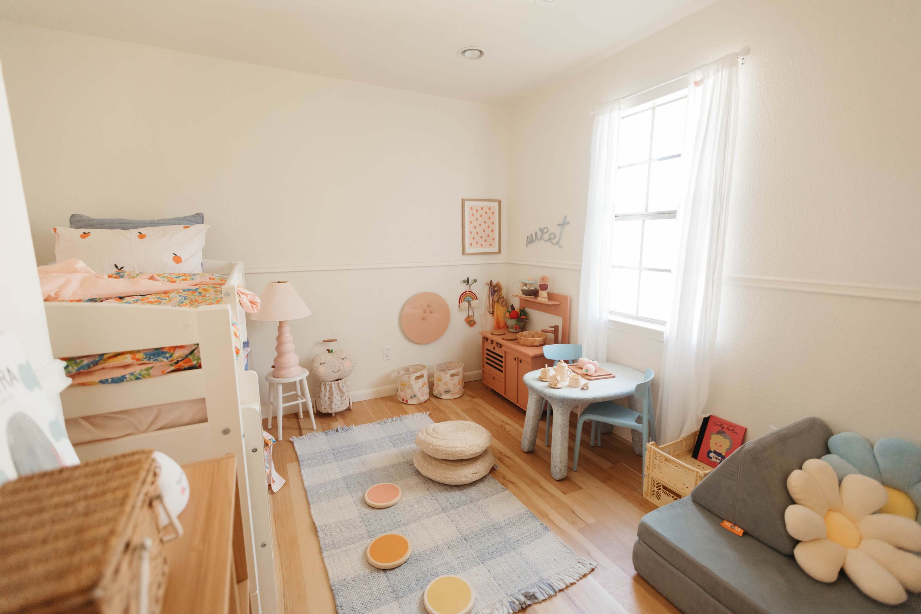 A brightly lit children's room with a loft bed, a soft rug, and a small table with chairs.