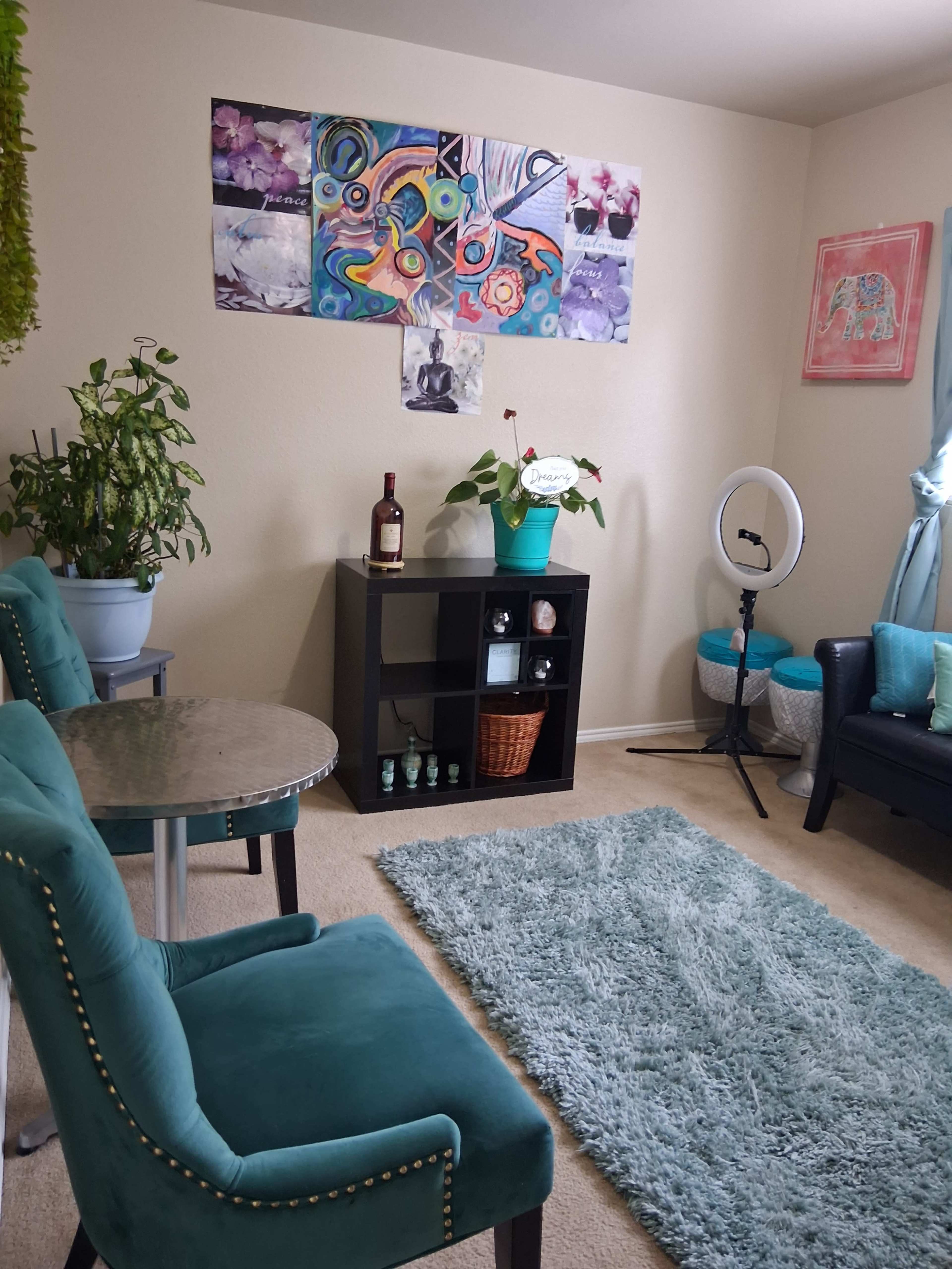 The image shows a cozy room with two teal chairs at a small table, a black bookshelf, a ring light, a rug, plants, and colorful wall art.