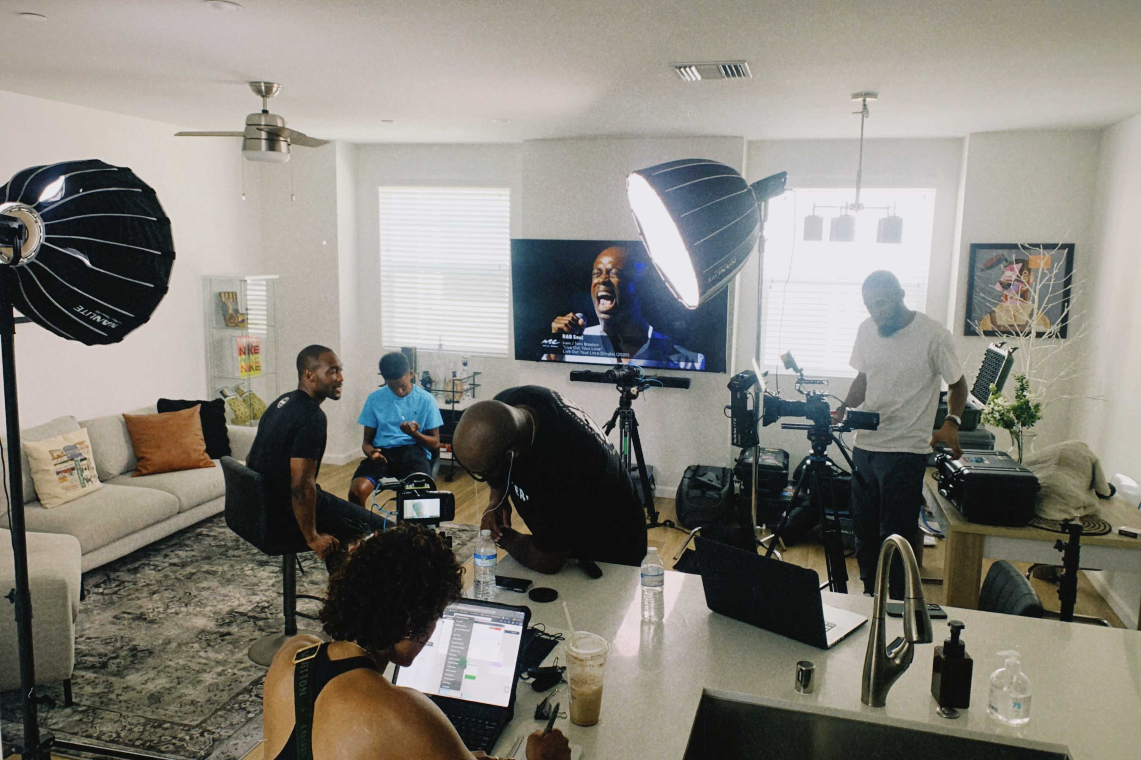 A group of five people work on laptops and set up filming equipment in a modern living room featuring a large TV displaying a performance.