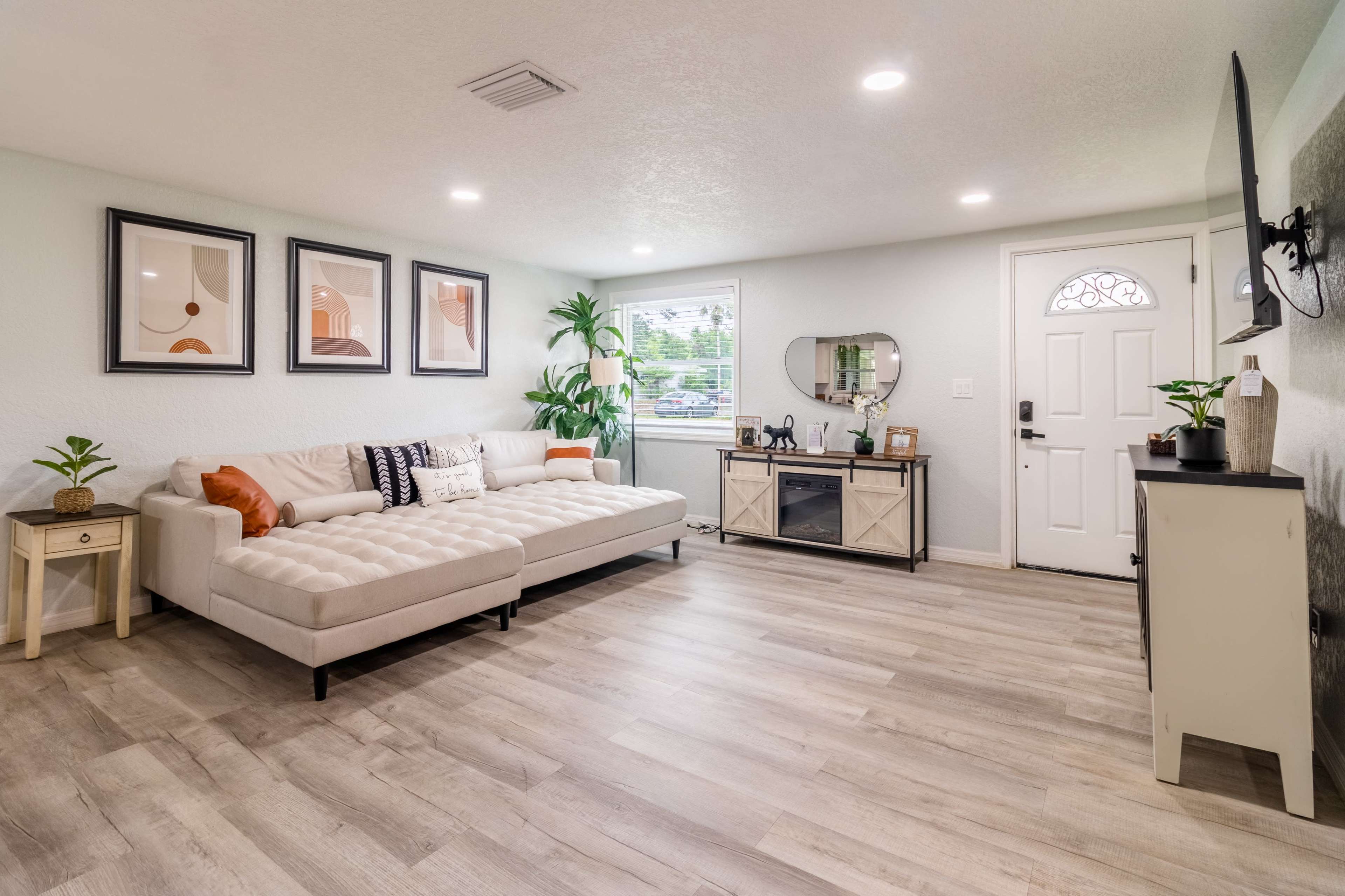 A light-colored living room features a sectional sofa, framed artwork on the walls, a media console, and a front door with a window.