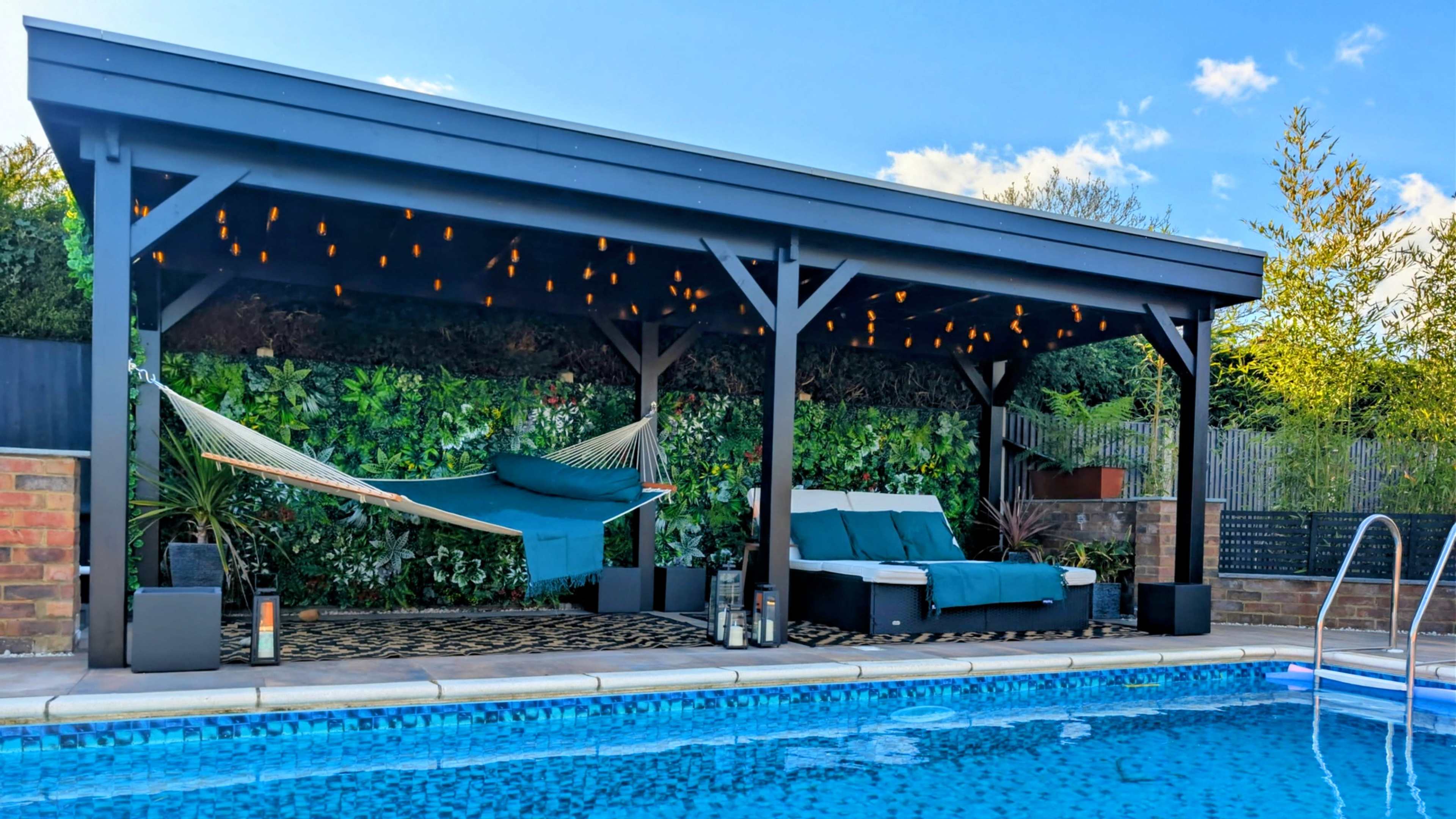 The image shows a covered seating area beside a swimming pool, featuring a hammock, lounge chairs, and string lights.