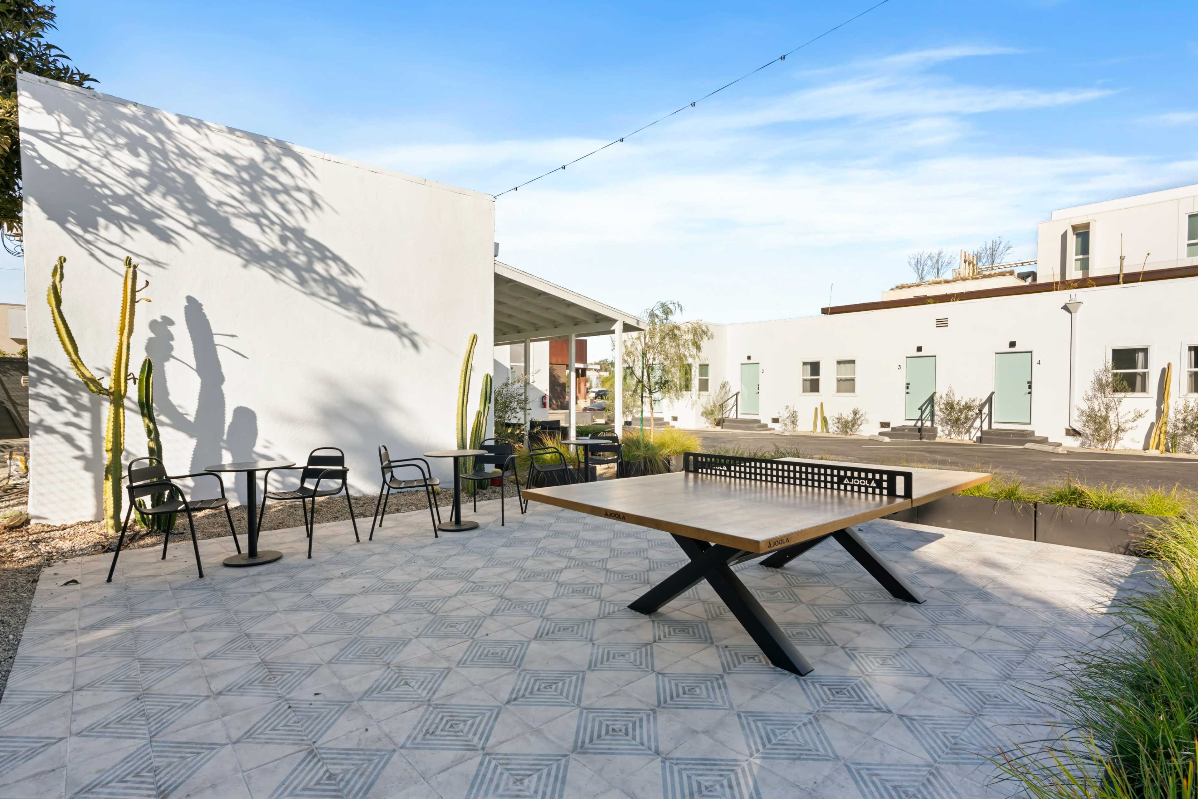 The image shows an outdoor area with a large table, chairs, and cacti, surrounded by white walls and buildings.