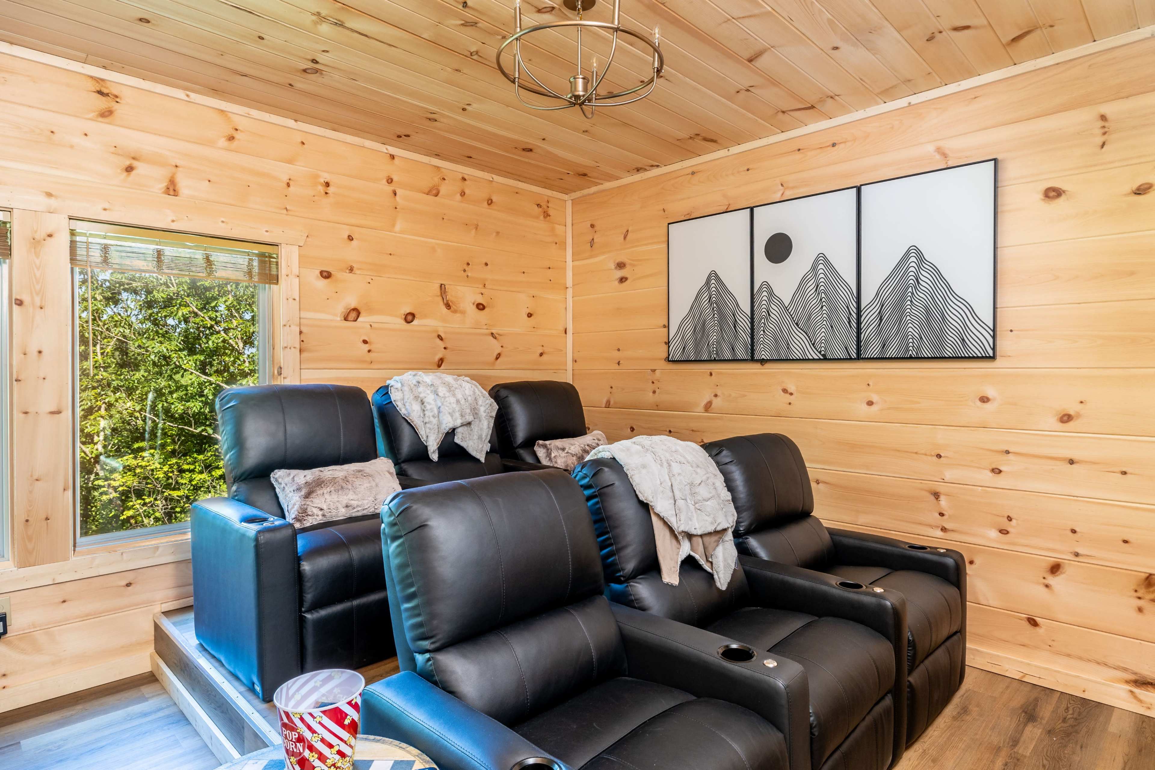 The image shows a cozy cabin-like living room featuring two black recliners with throw blankets, a large window with green views outside, and a modern mountain-themed artwork on the wall.