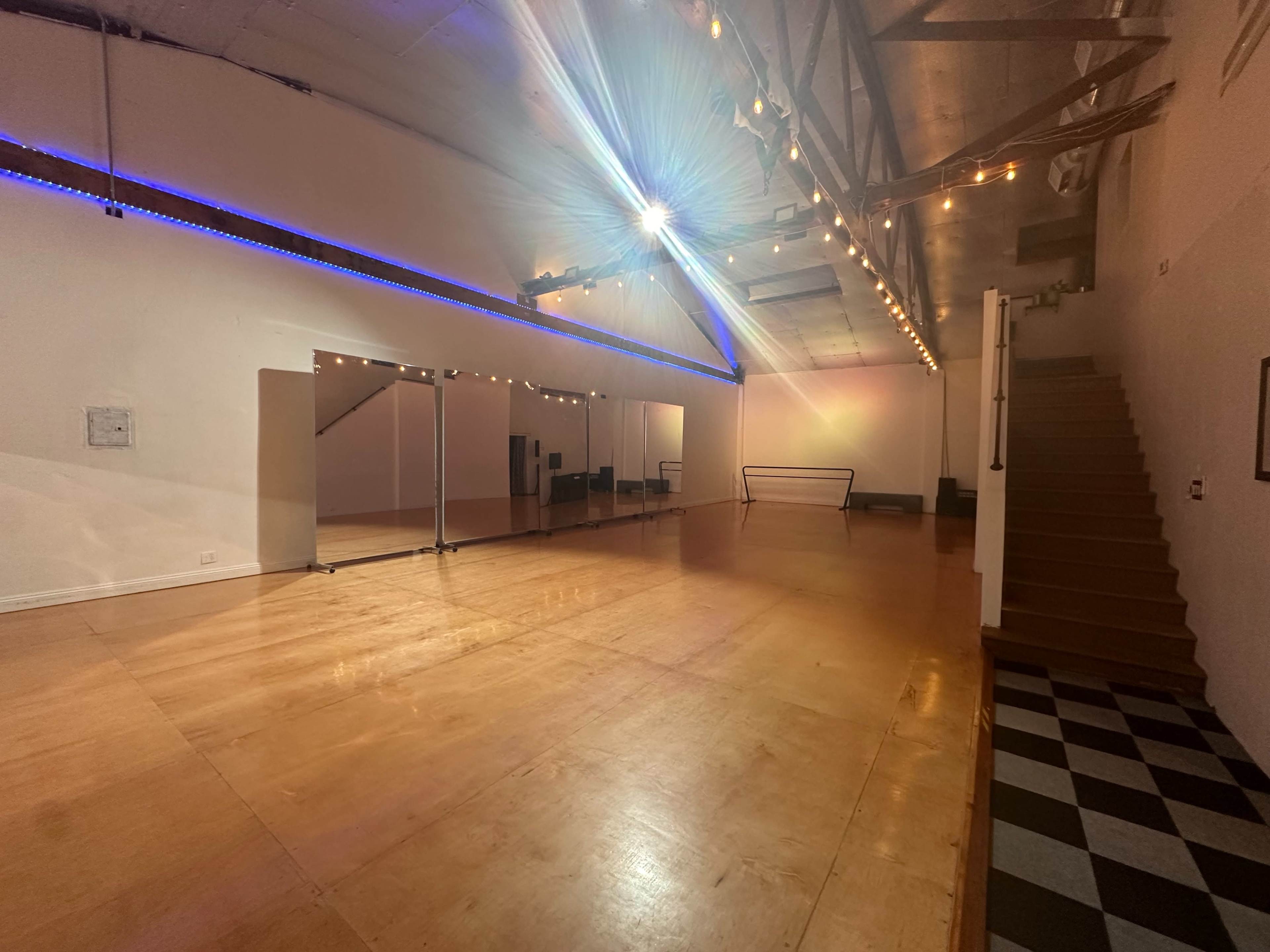 The image shows a spacious empty dance studio with wooden flooring, large mirrors on one wall, and stair access to a loft area.