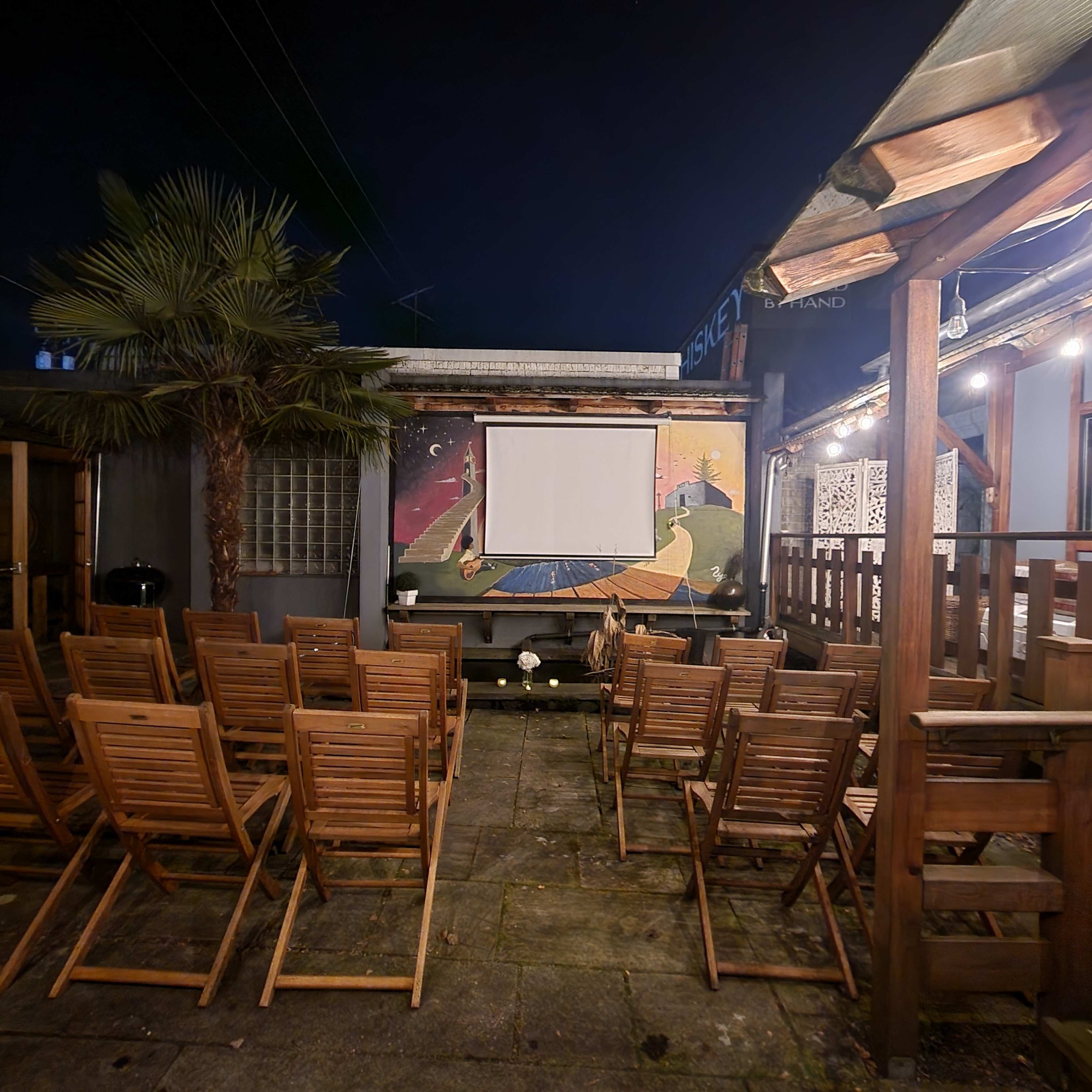 The outdoor seating area is prepared for a screening, featuring wooden chairs arranged in front of a blank projector screen with a colorful mural in the background.