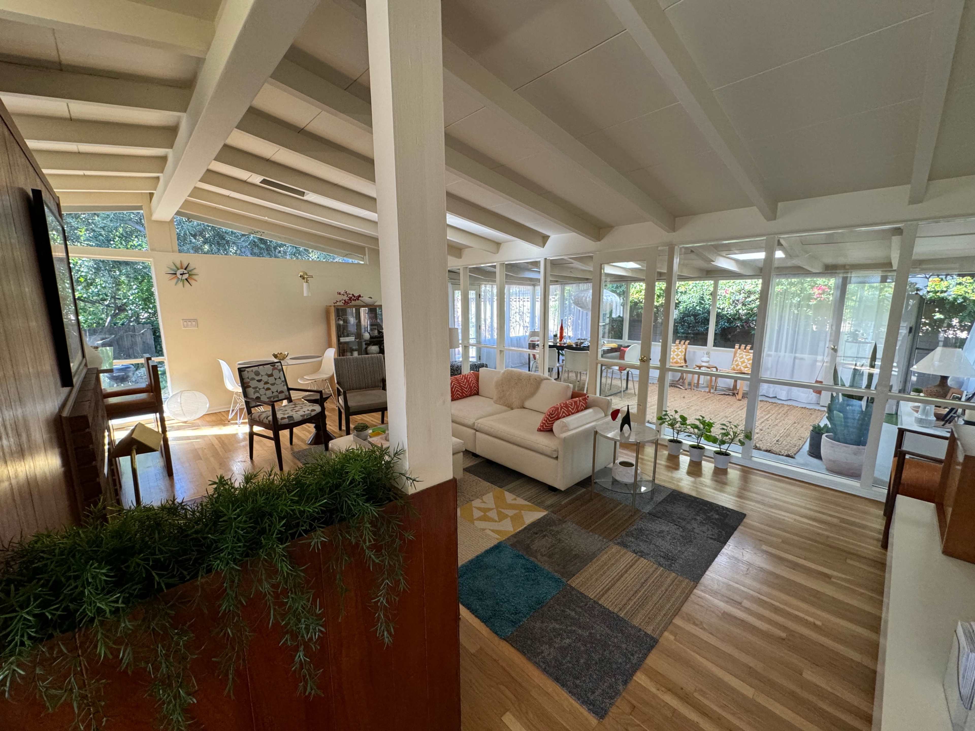 The image shows a bright and airy living room with vaulted ceilings, hardwood floors, and large glass windows providing a view of a garden.