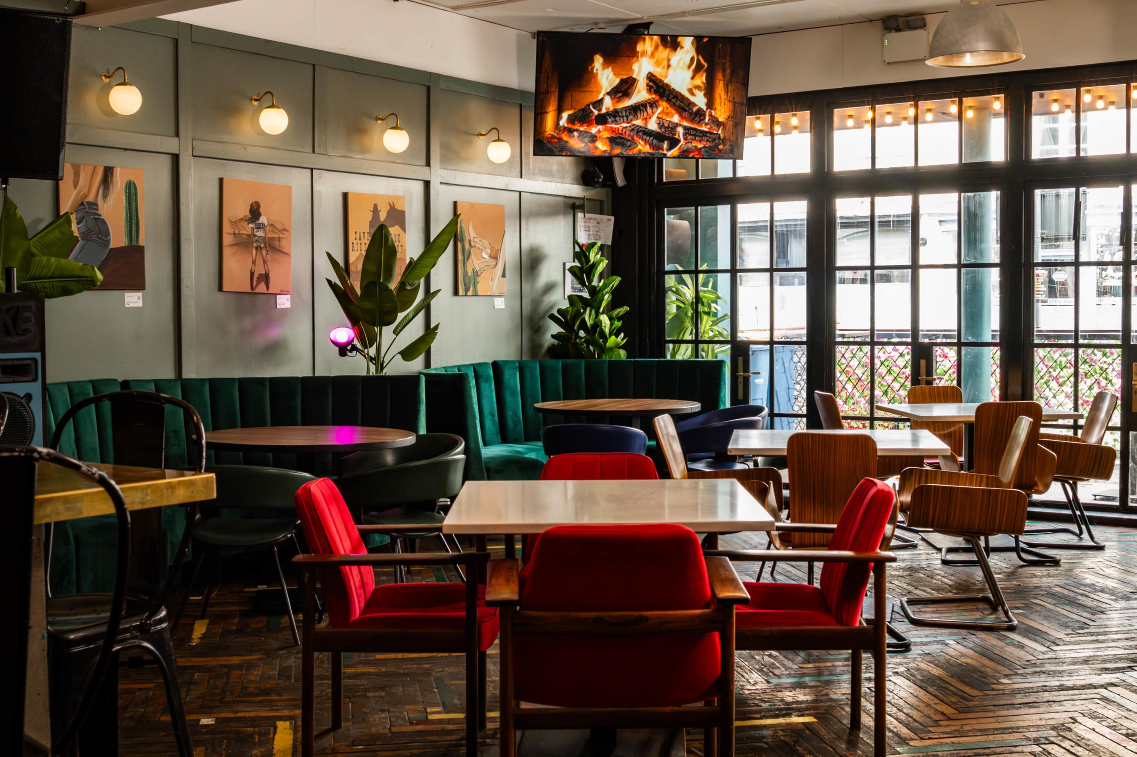 The image shows a cozy café interior featuring green velvet seating, wooden tables, and a mounted screen displaying a fireplace, with large windows allowing natural light to enter.