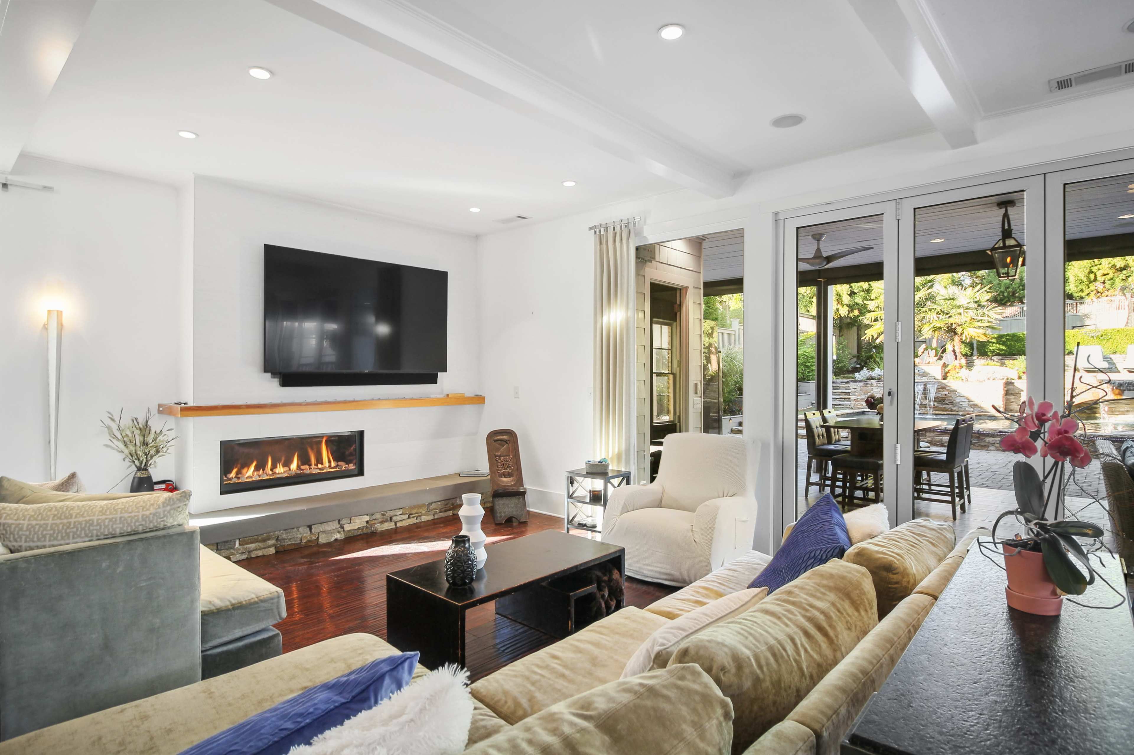 A modern living room with a flat-screen TV mounted above a fireplace, featuring comfortable seating, decorative plants, and large glass doors leading to an outdoor space.