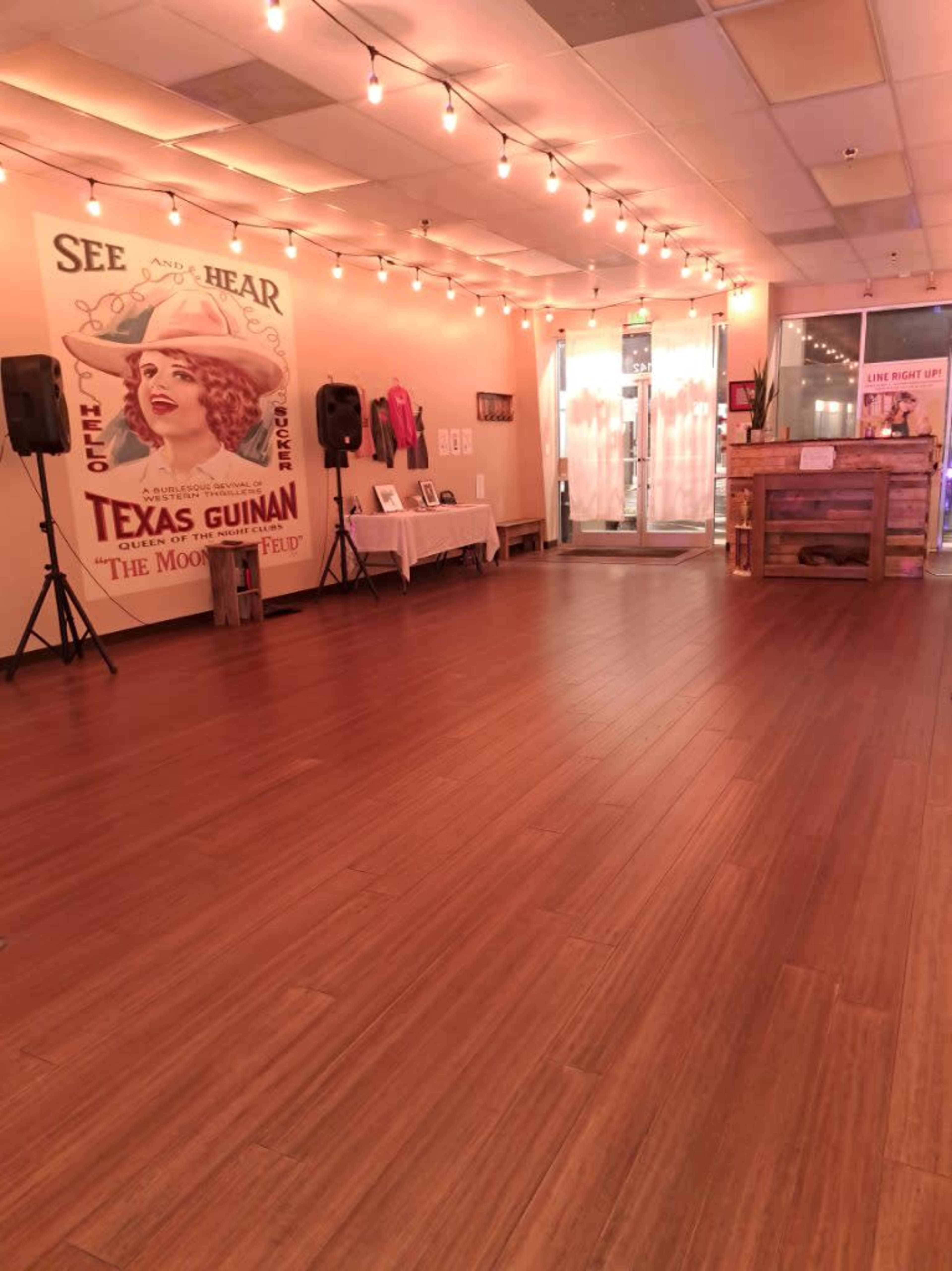 The interior of a spacious venue features wooden flooring, string lights overhead, a vintage poster on the wall, and a welcoming entrance area.