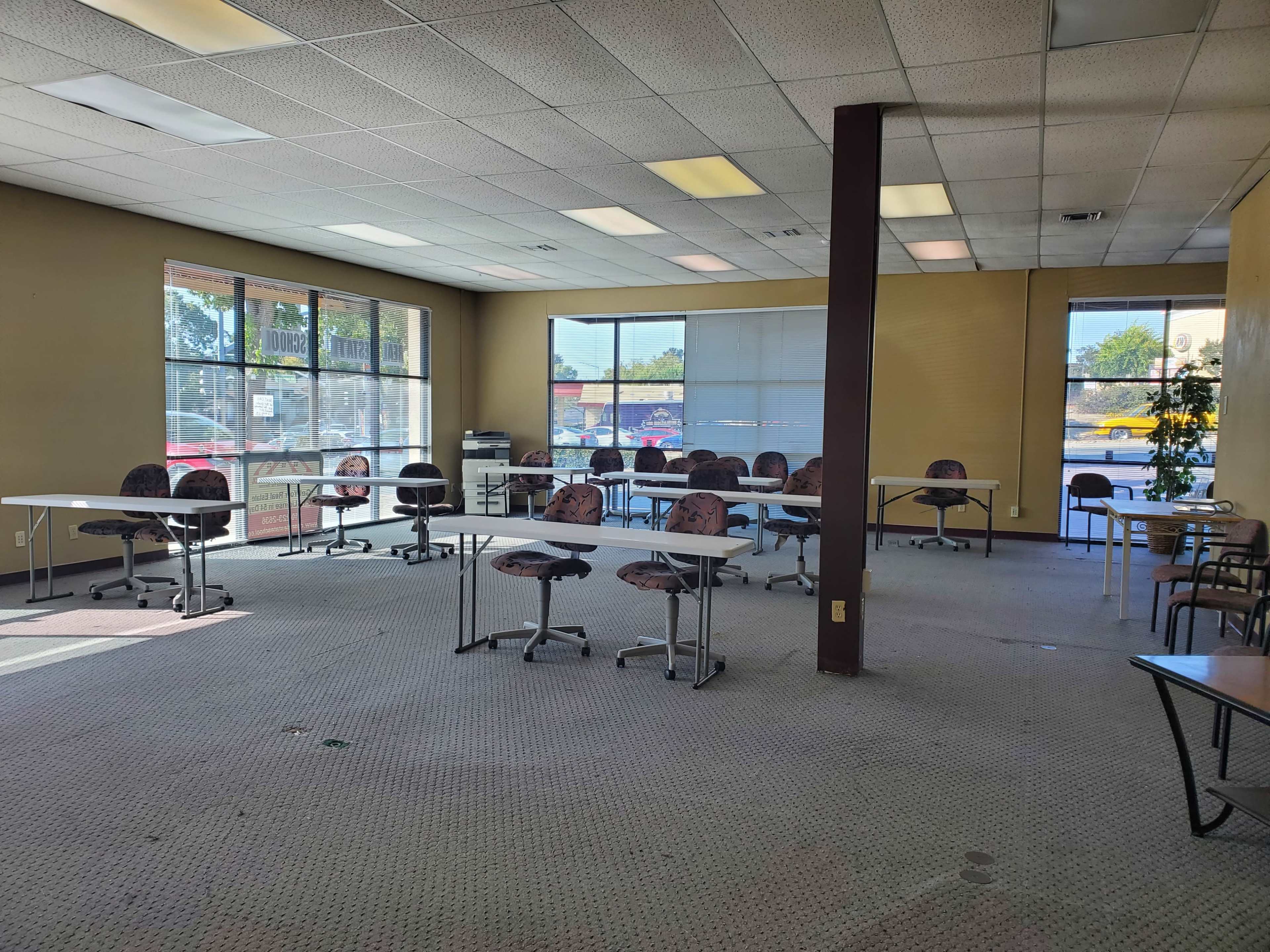 An empty classroom with several tables and chairs arranged in rows, illuminated by natural light from large windows.