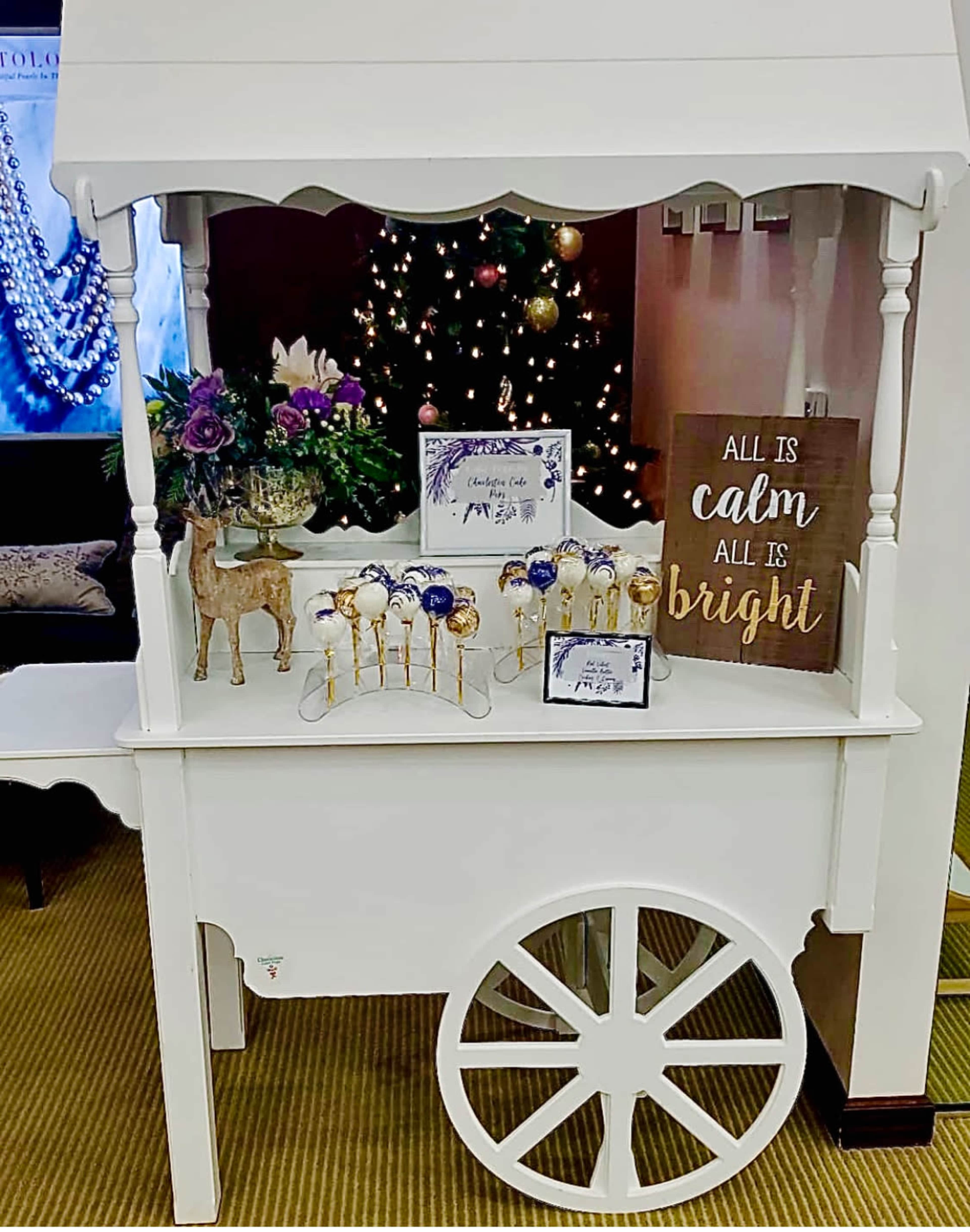 A white cart displays decorative lollipops, a wooden sign with a festive message, and floral arrangements in front of a Christmas tree.