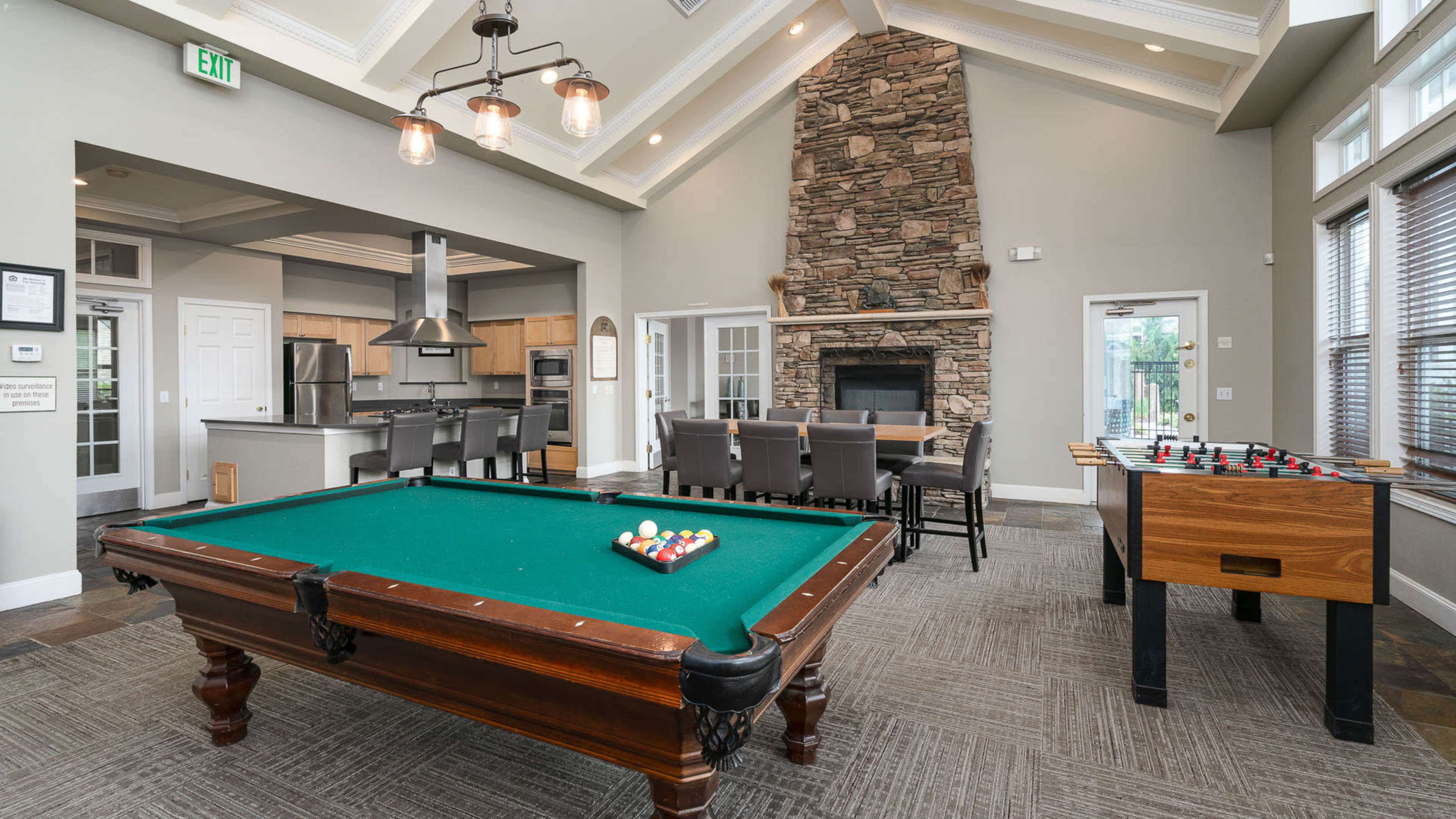 The image shows a communal recreation area featuring a pool table, foosball table, a stone fireplace, and dining seating situated near a kitchen.