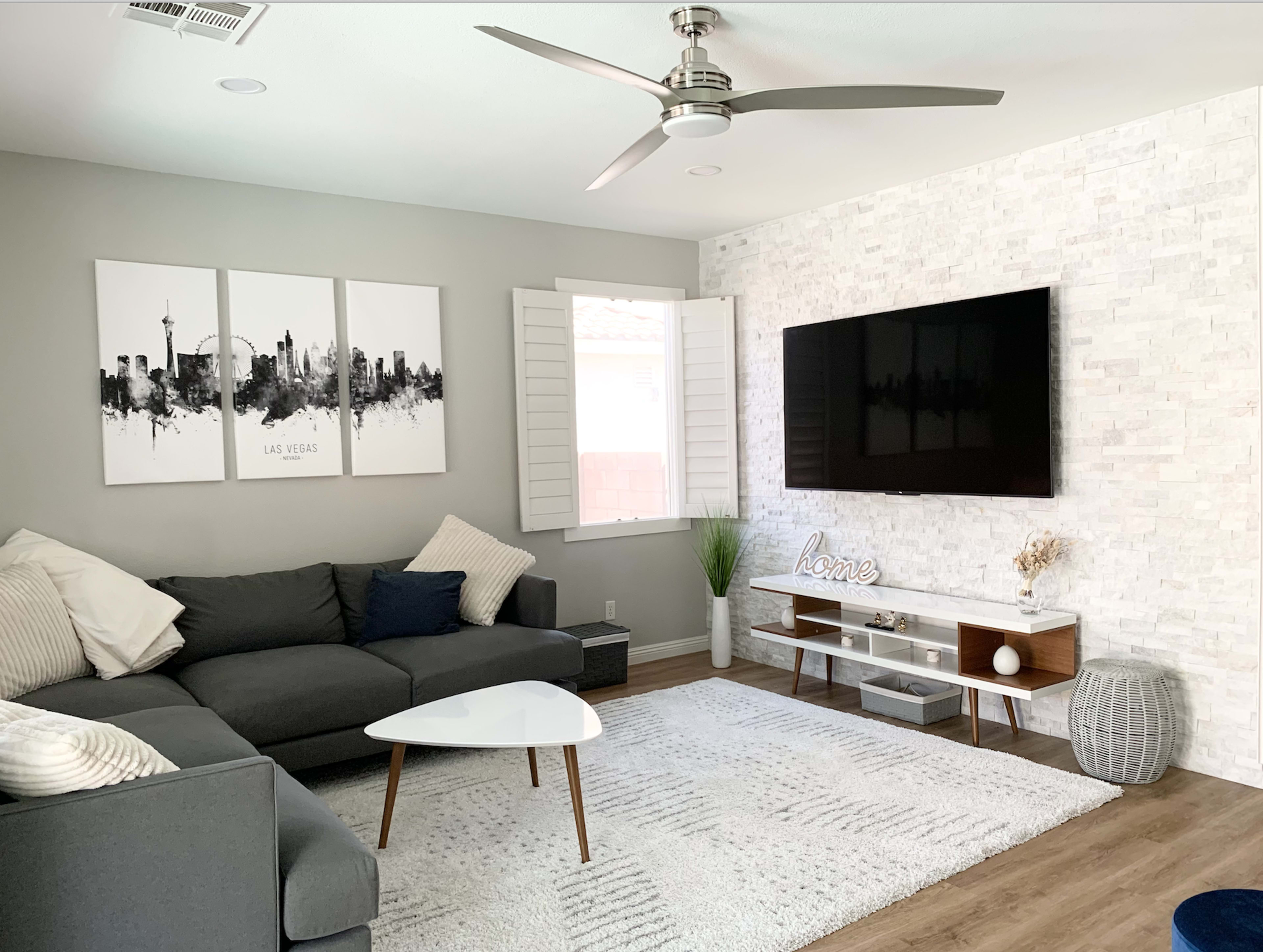 The living room features a gray sectional sofa, a wall-mounted television, and a decorative area with framed artwork and potted plants.