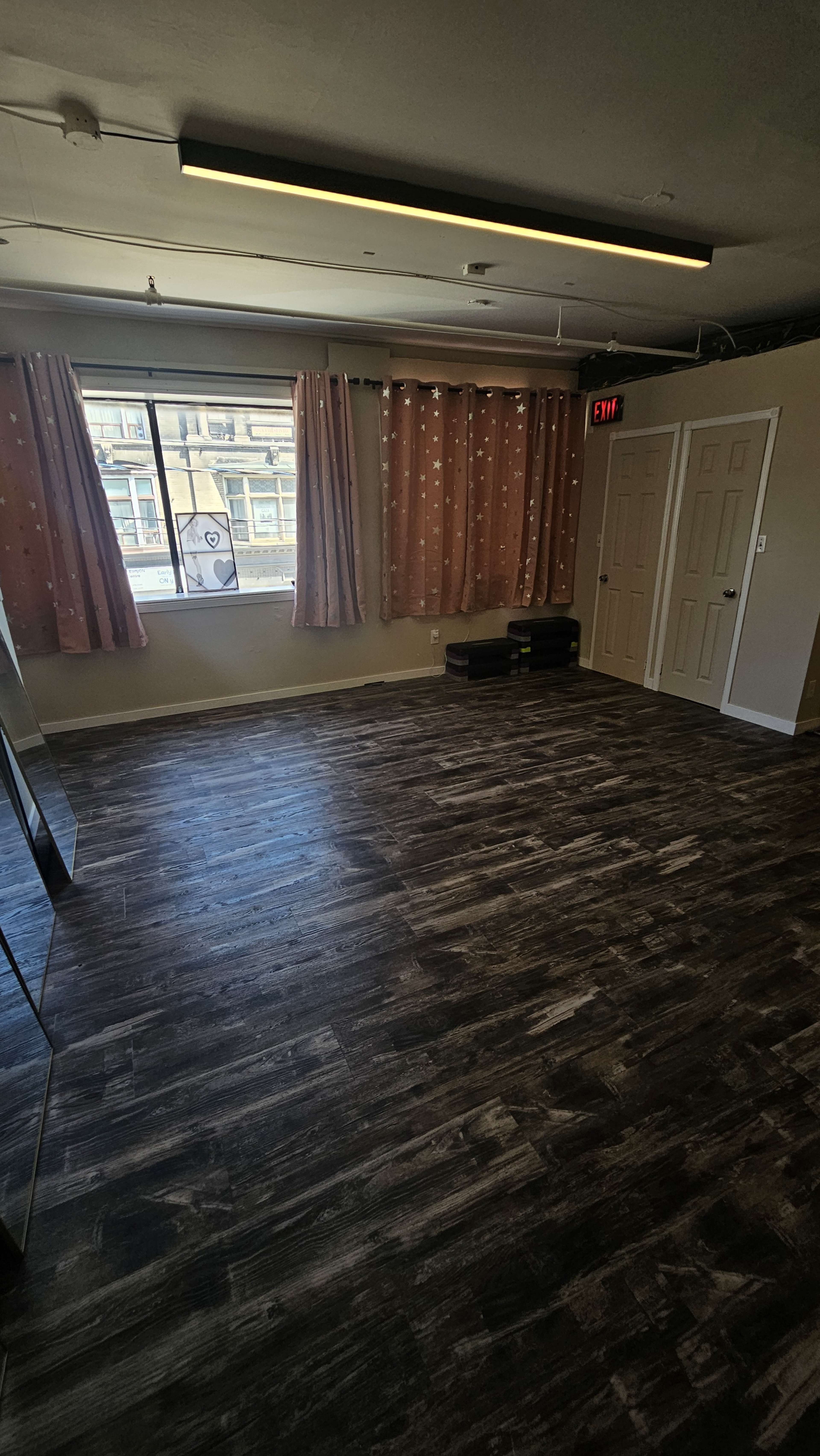 The image shows an empty room with wooden flooring, a large window with curtains, and two closed doors.