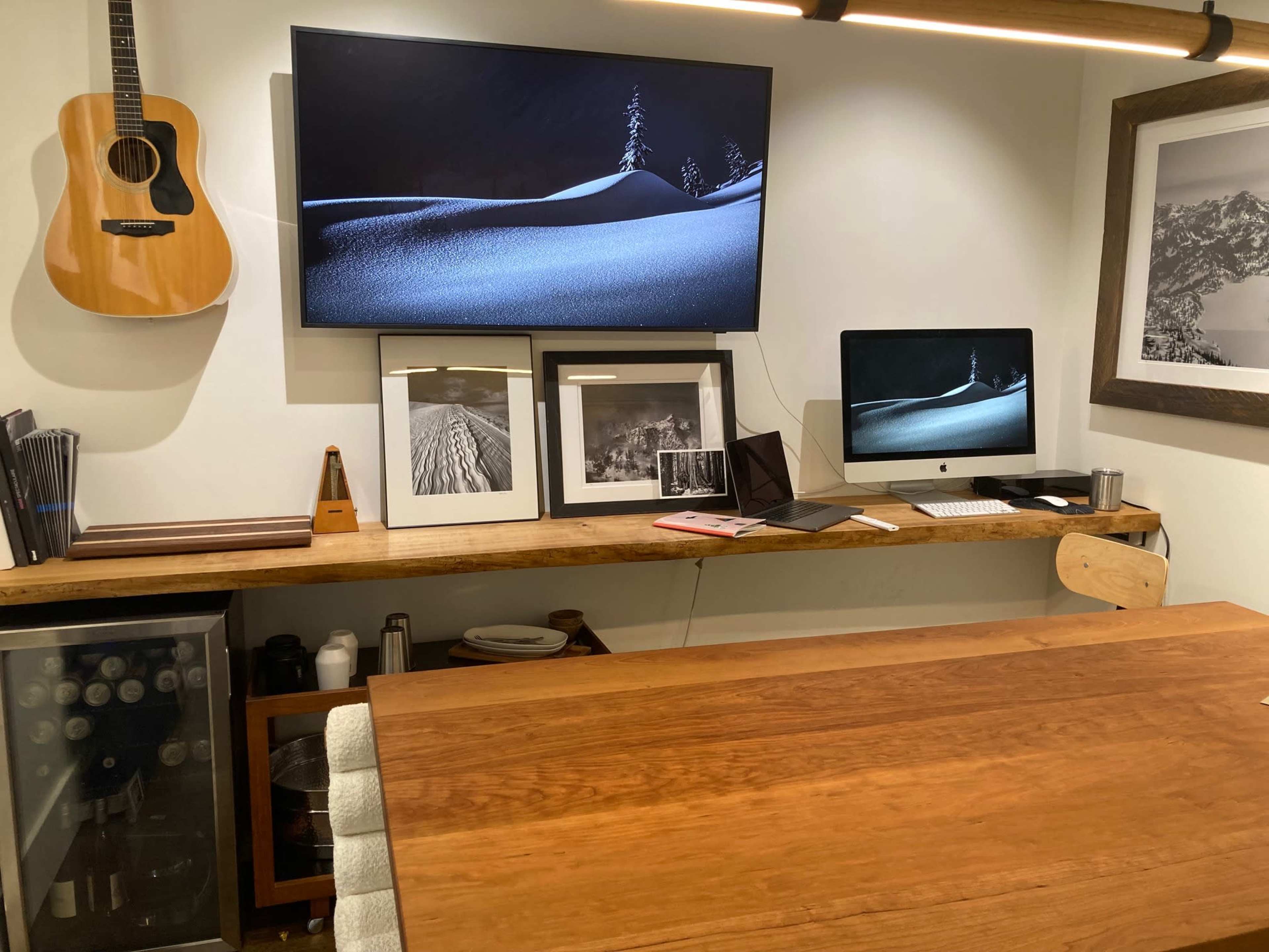 A workspace featuring a wooden table, a wall-mounted TV, a guitar, framed photographs, and a desktop computer on a wooden shelf.