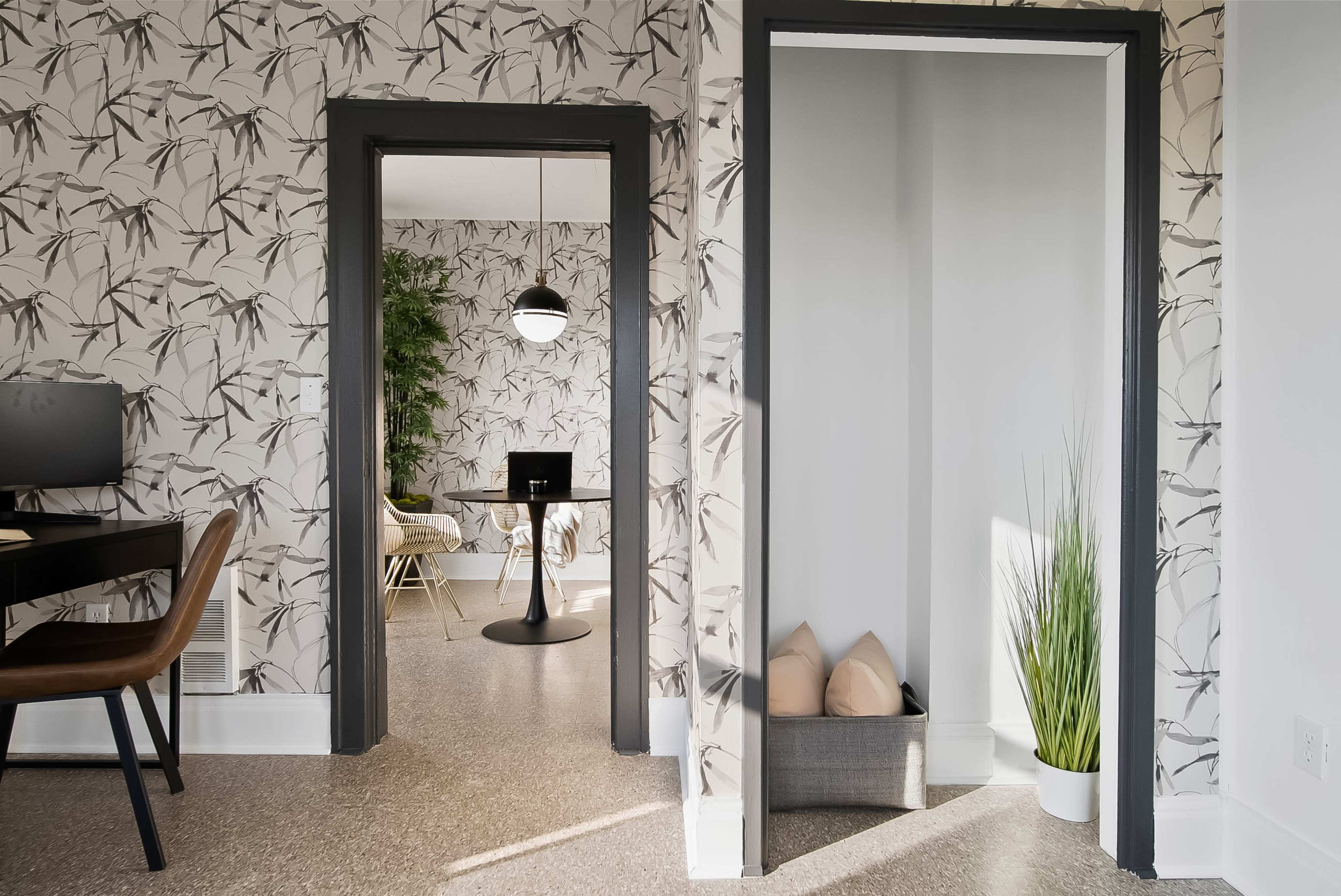A view of a brightly lit room featuring patterned wallpaper, with a doorway leading to a second room that includes a round table and a potted plant.