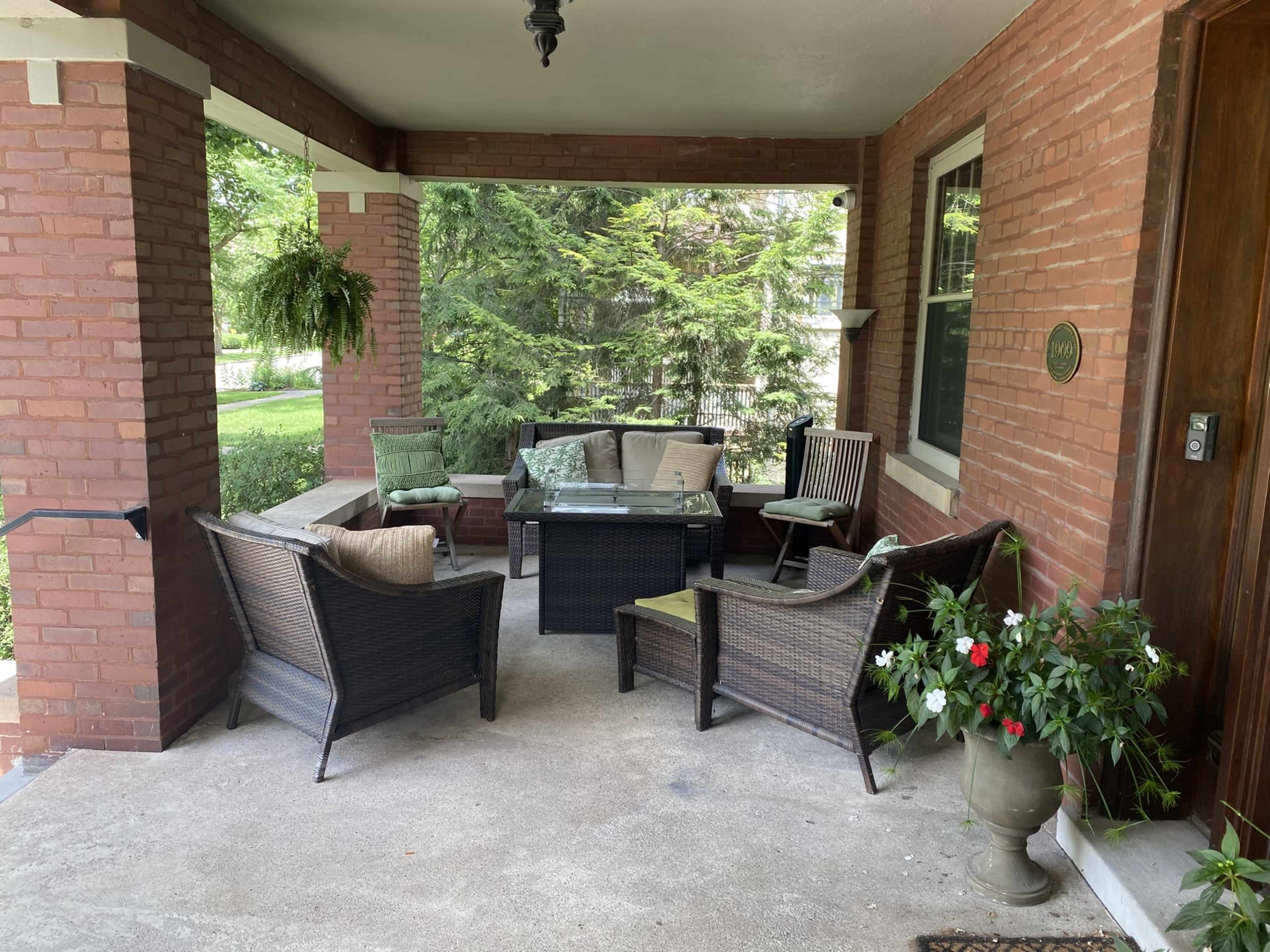 The image shows a porch with a set of wicker furniture, including a coffee table and several chairs, surrounded by greenery.