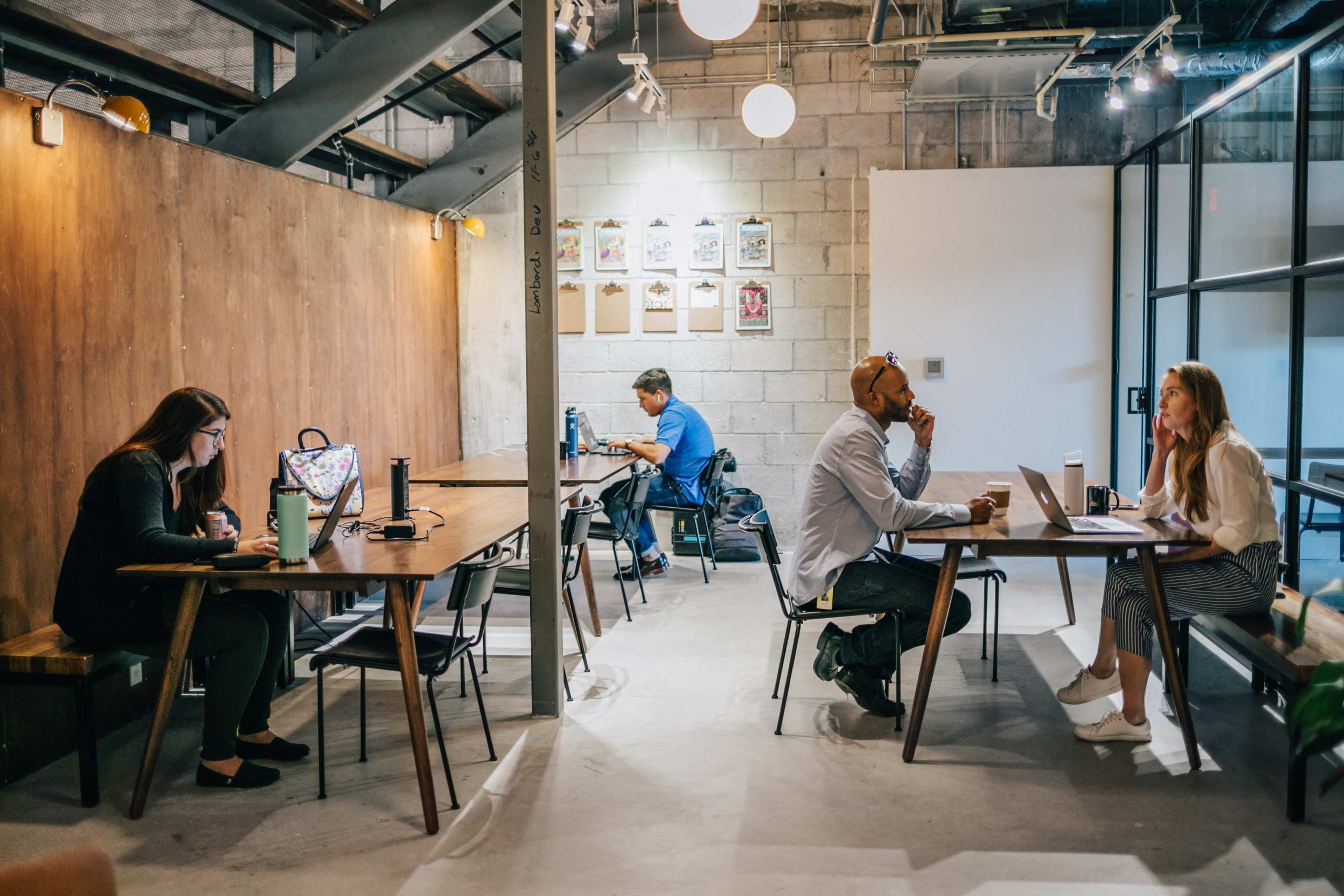 A modern office setting with four individuals working at different tables in a well-lit space featuring exposed brick and wooden accents.