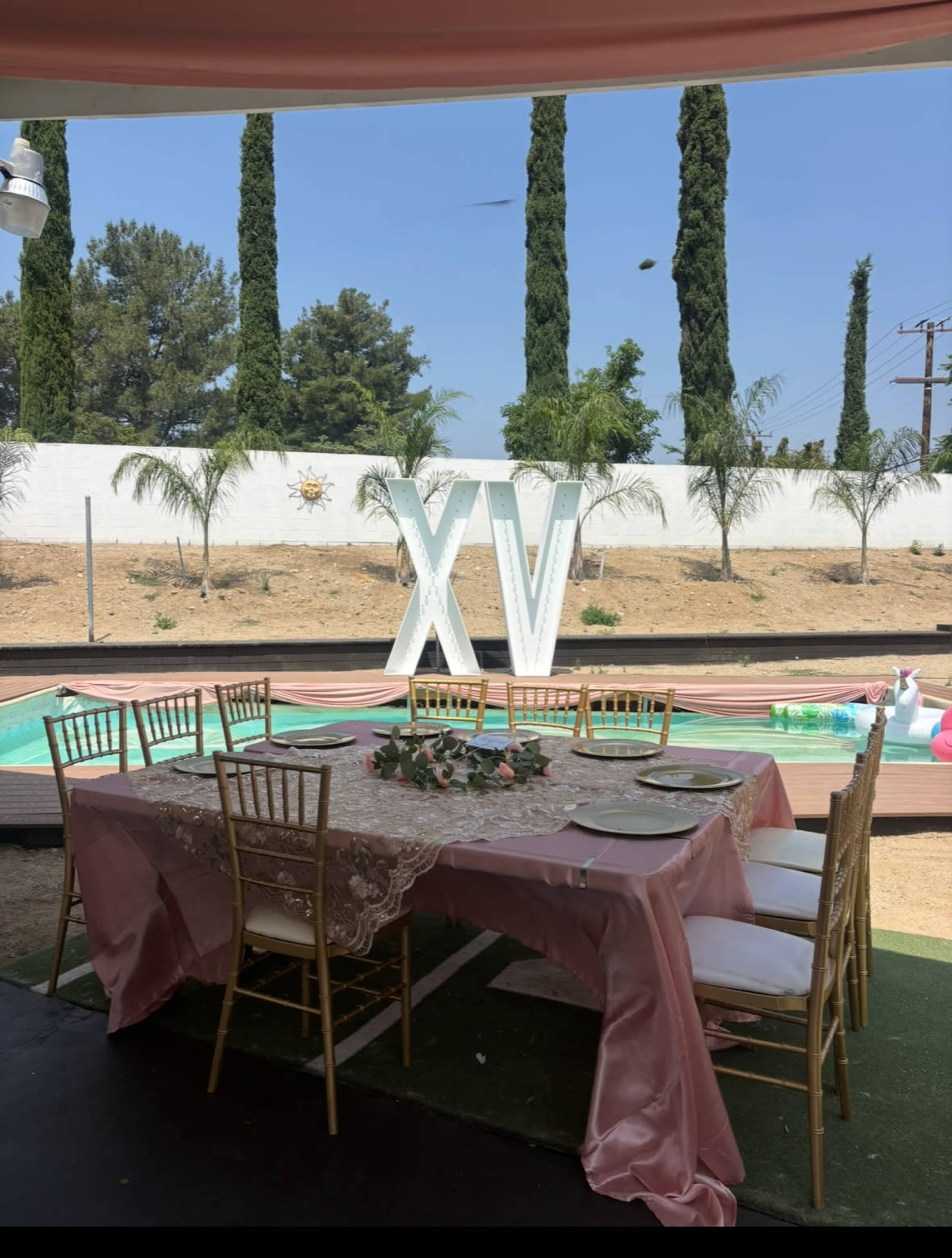 A decorated dining area with a table set for a celebration is positioned by a pool, featuring large "XV" letters in the background.