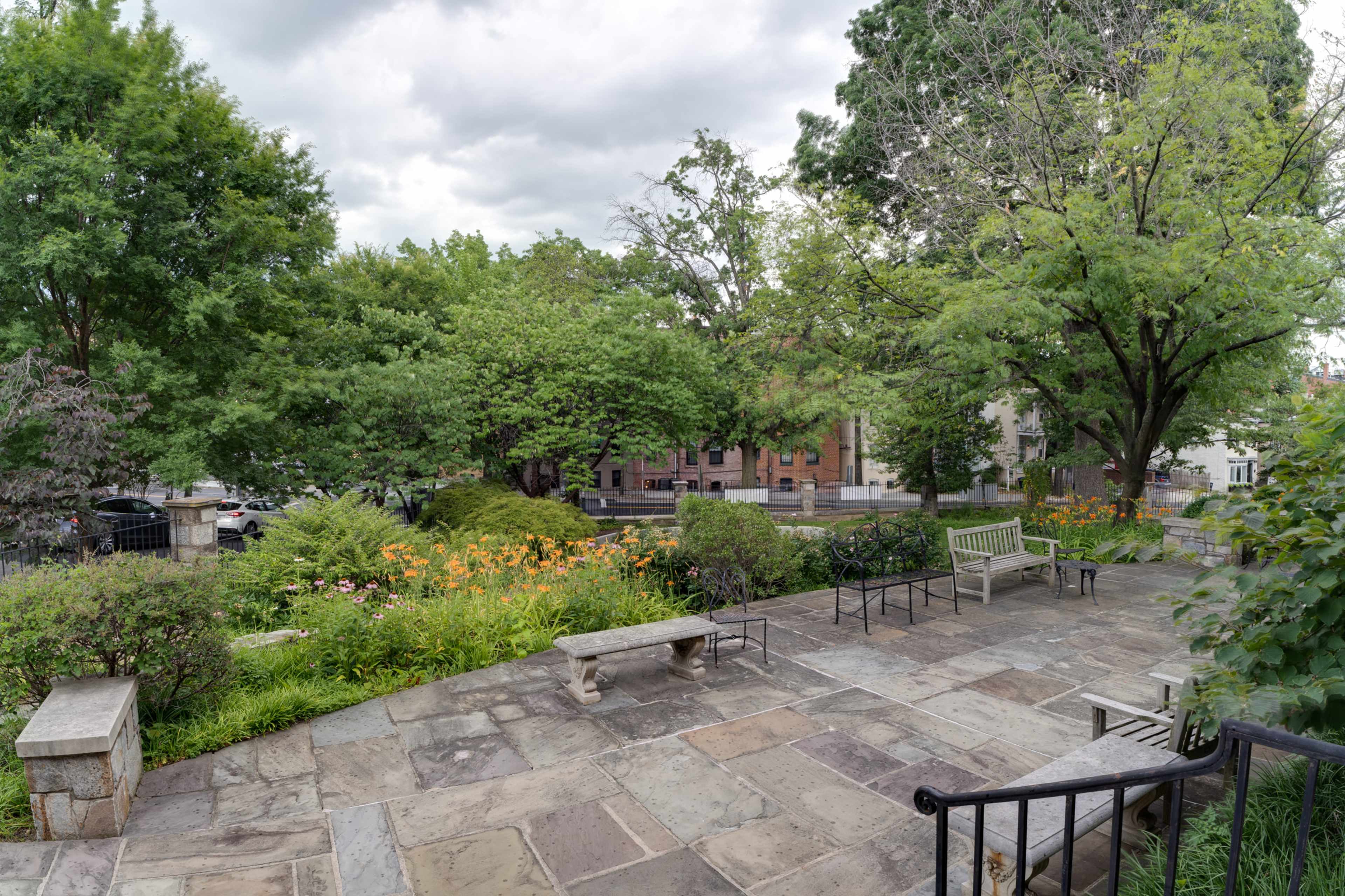 A stone patio with benches overlooks a garden filled with green trees and colorful flowers.