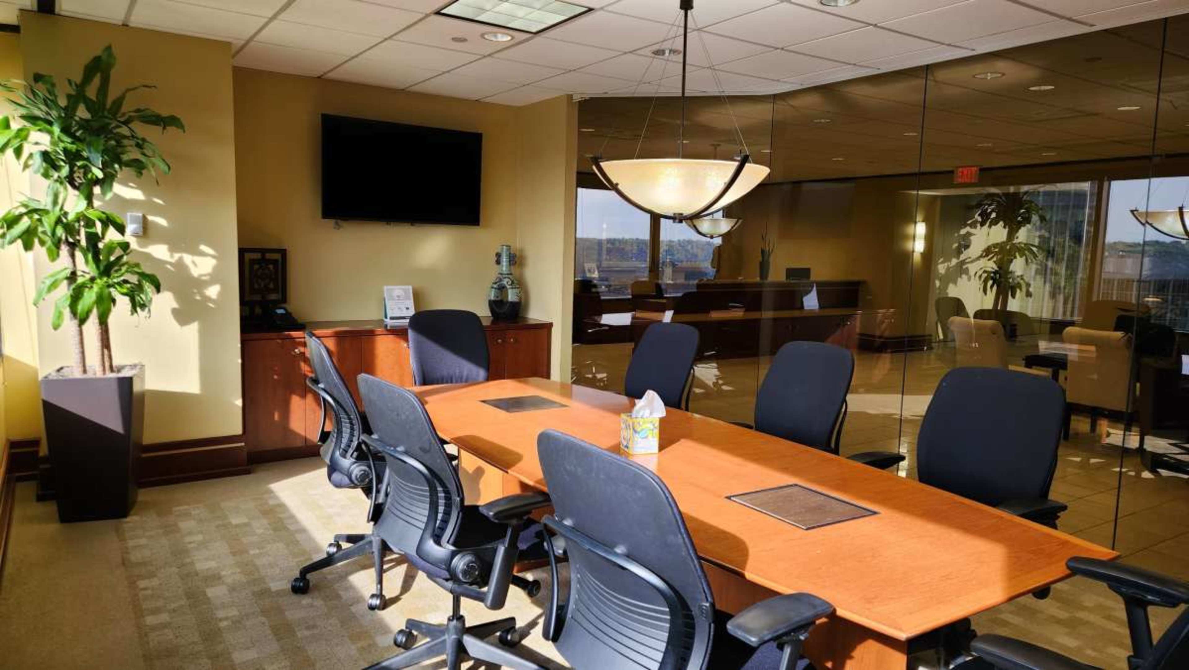A conference room features a long wooden table surrounded by black office chairs, with a large window offering a view outside and a television mounted on the wall.