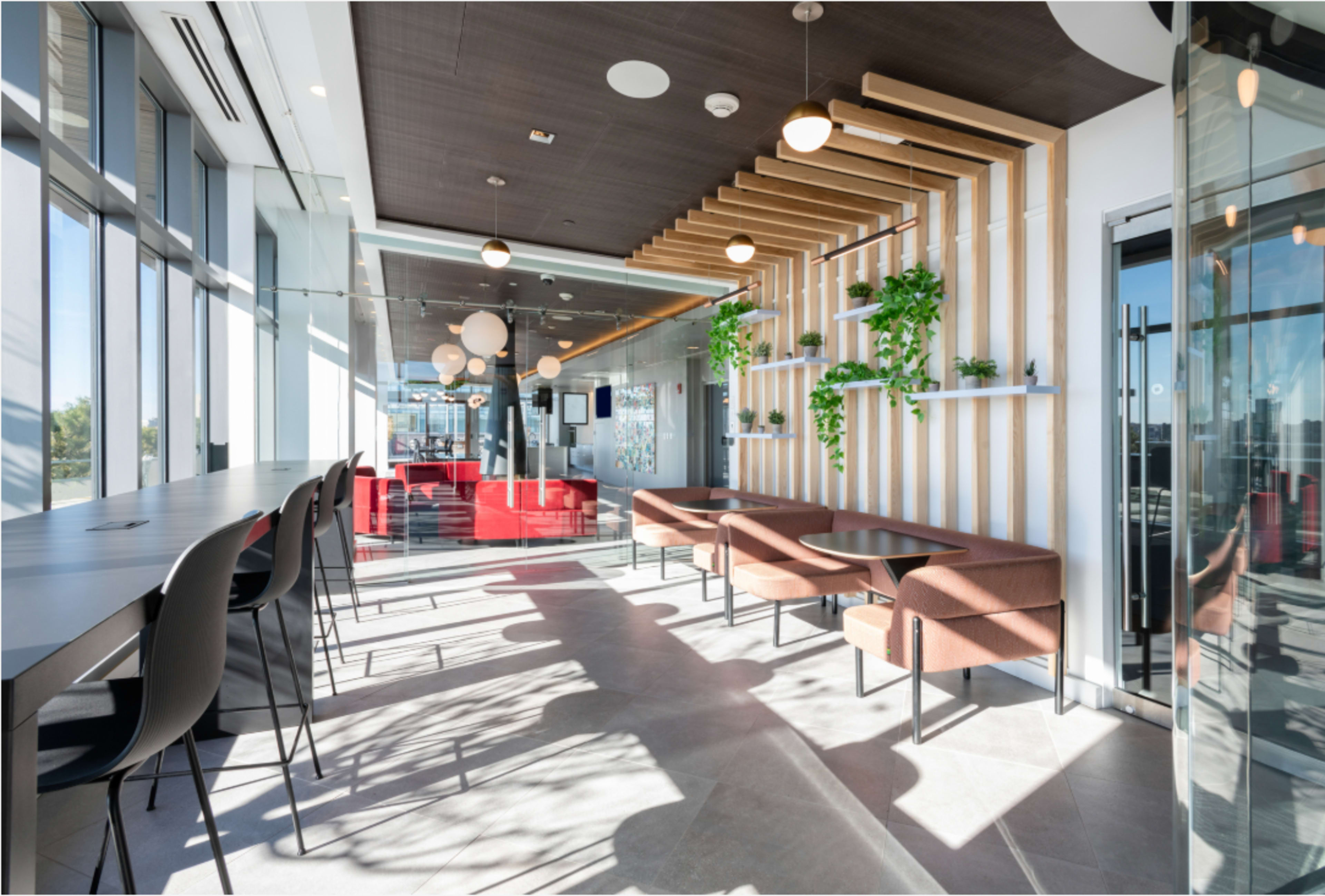 The image shows a modern, well-lit lounge area featuring sleek furniture, large windows, and decorative plants along a wooden feature wall.