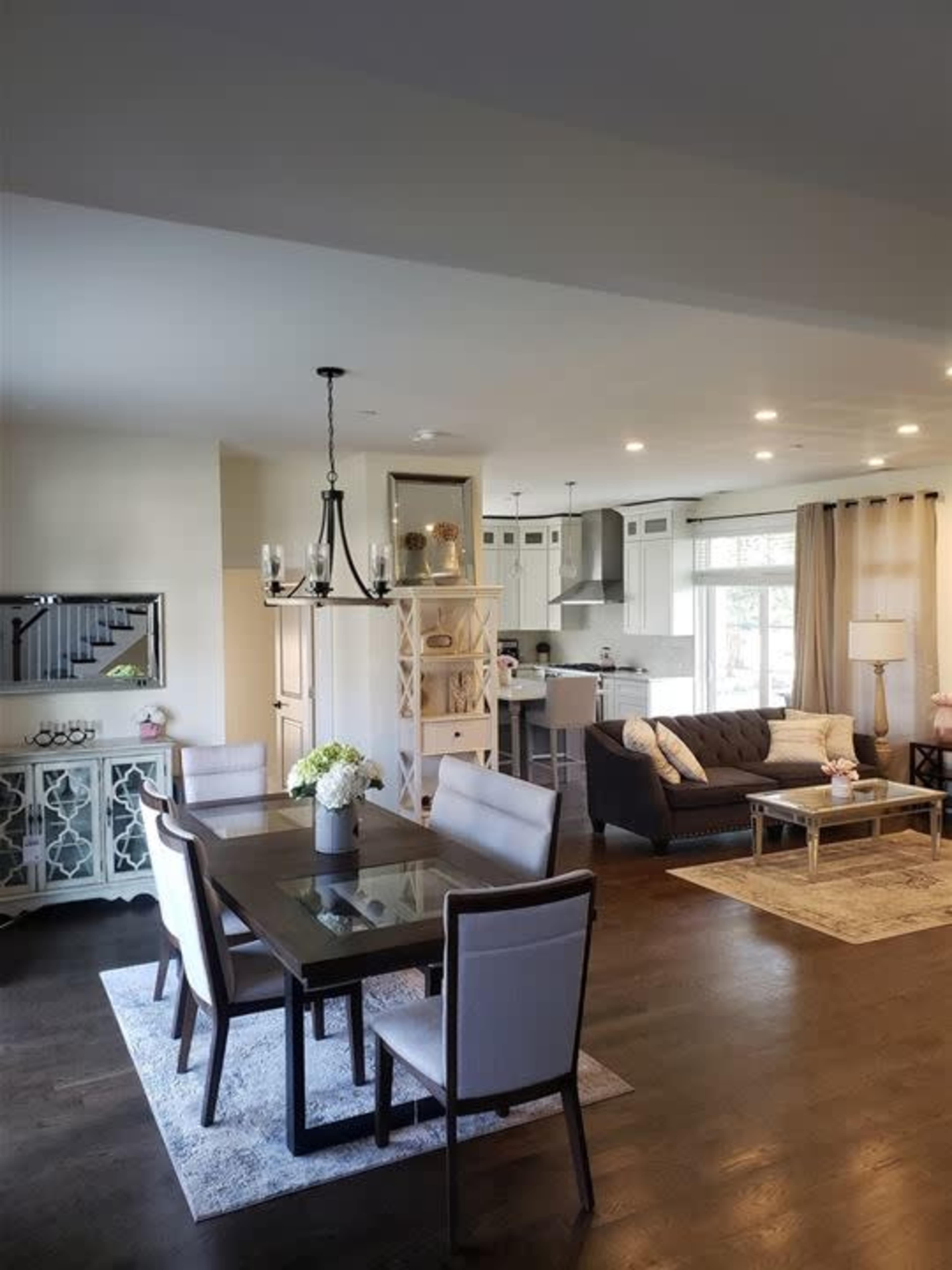 The image shows a modern living and dining area with a glass dining table, gray chairs, a brown sofa, and a light-colored kitchen in the background.