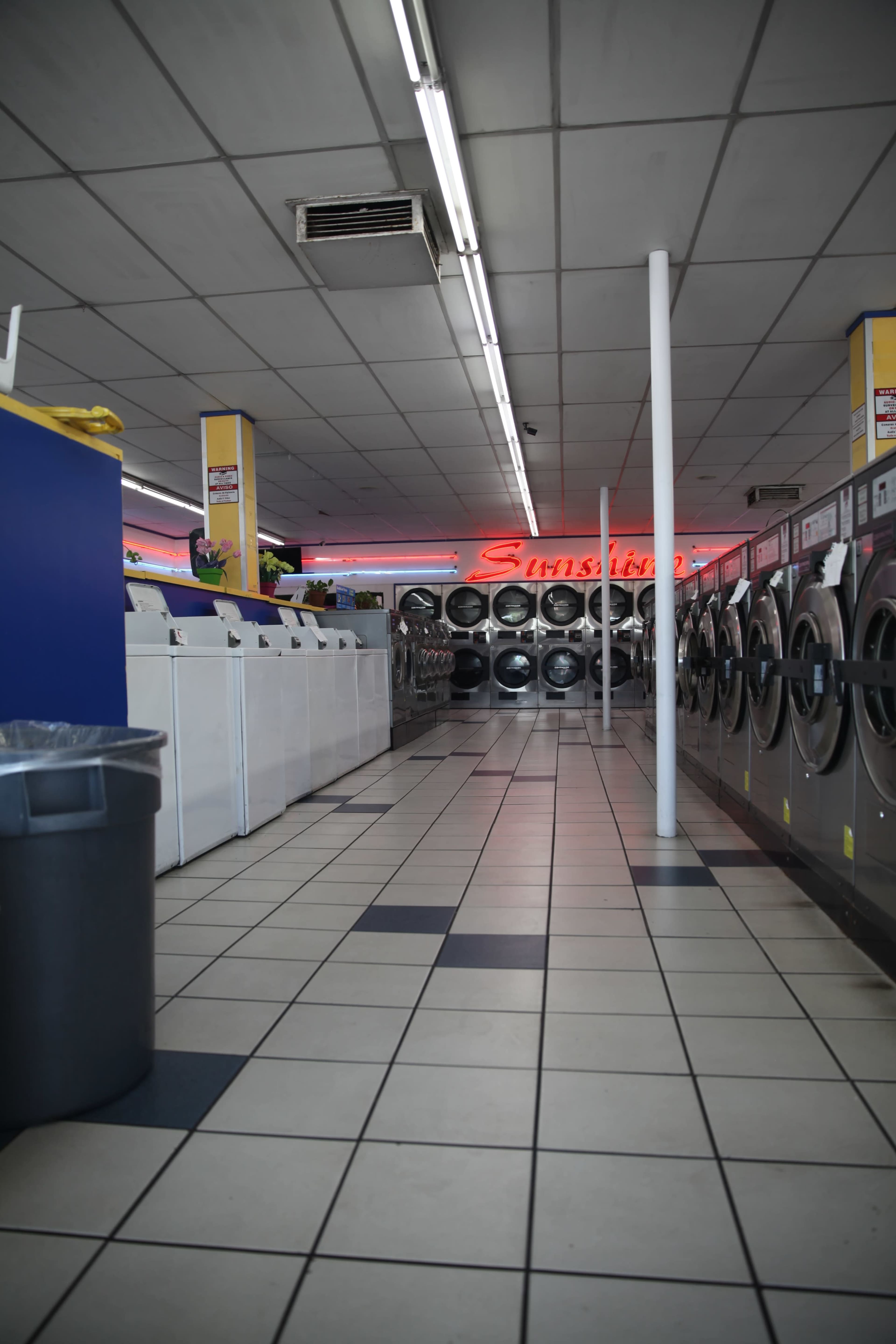 A laundromat features rows of washing machines and dryers, with a central aisle leading to a neon sign reading "Sunshine."