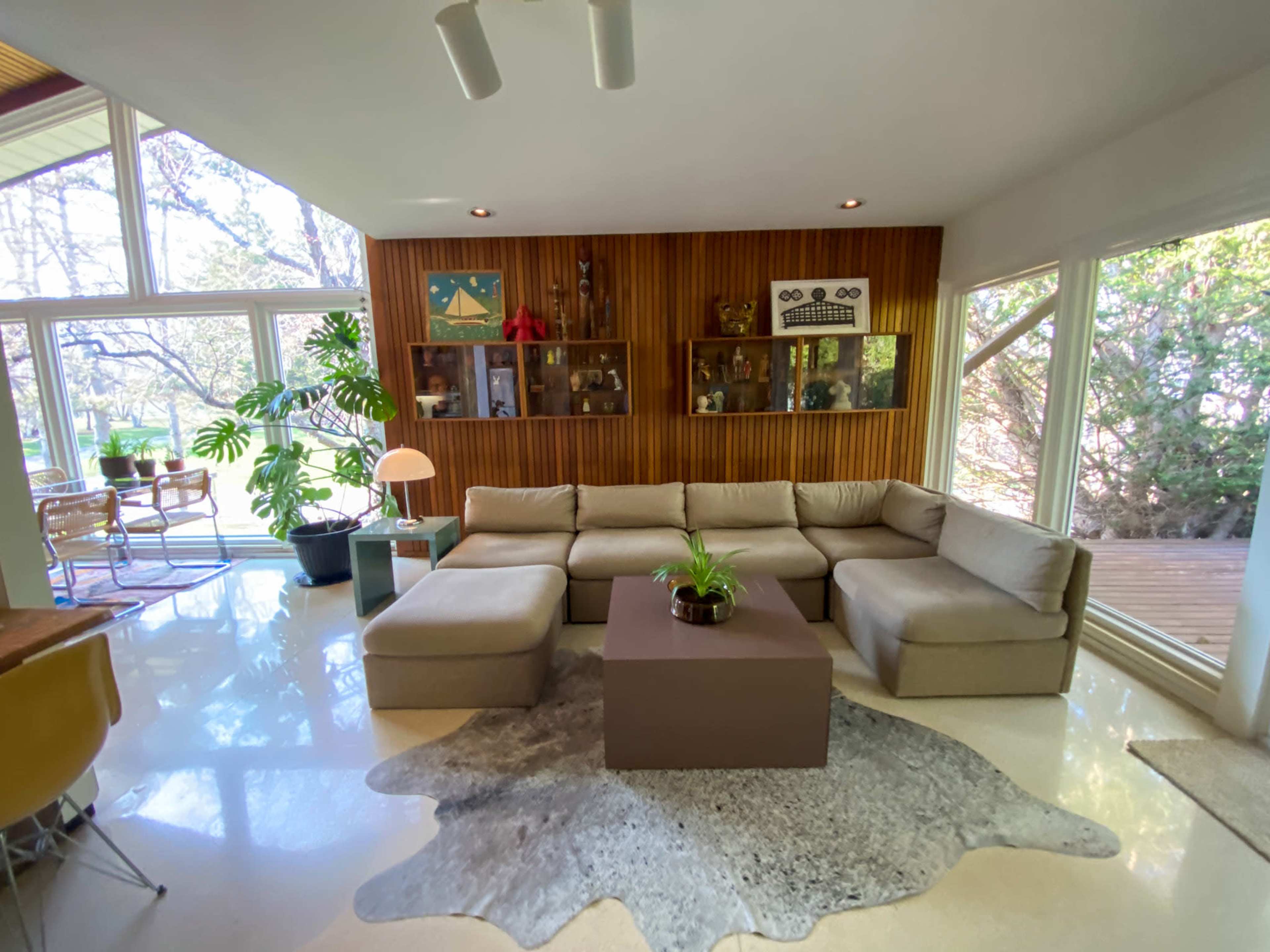 The image shows a modern living room with a large sectional sofa, a central coffee table, and large windows that provide natural light and a view of greenery outside.