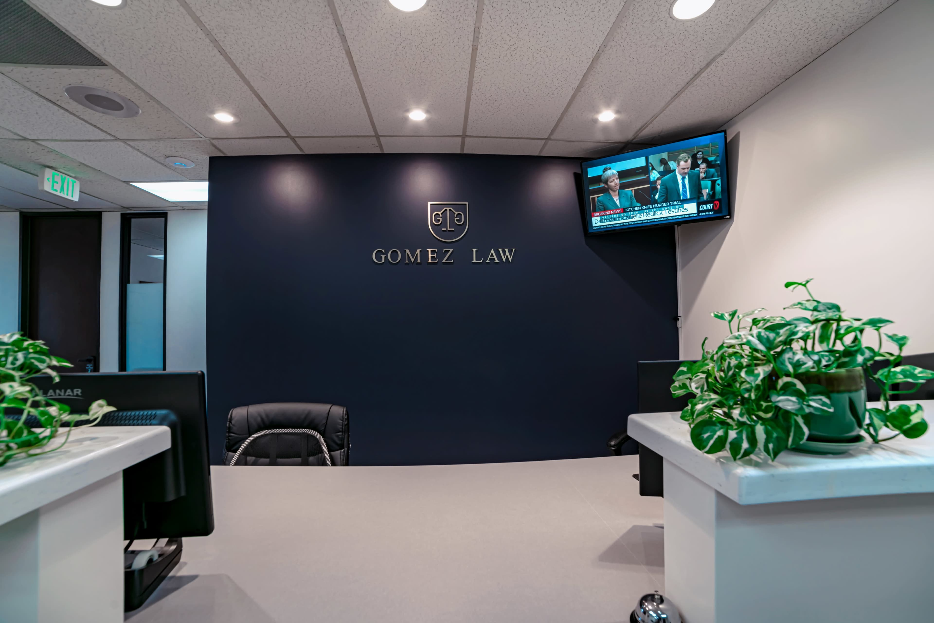 A law office reception area with a dark blue wall featuring the logo "GOMEZ LAW," a television displaying news, and potted plants on the reception desk.